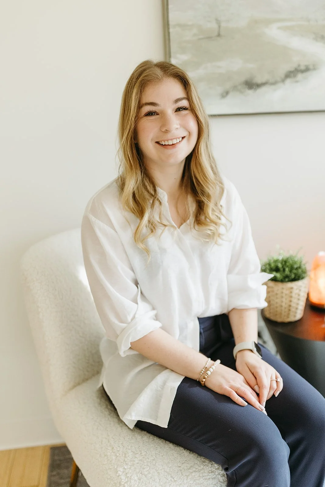 A young woman with long red hair sitting on a white textured chair, smiling, wearing a white blouse and dark pants, with a potted plant and salt lamp on a nearby table in the background.