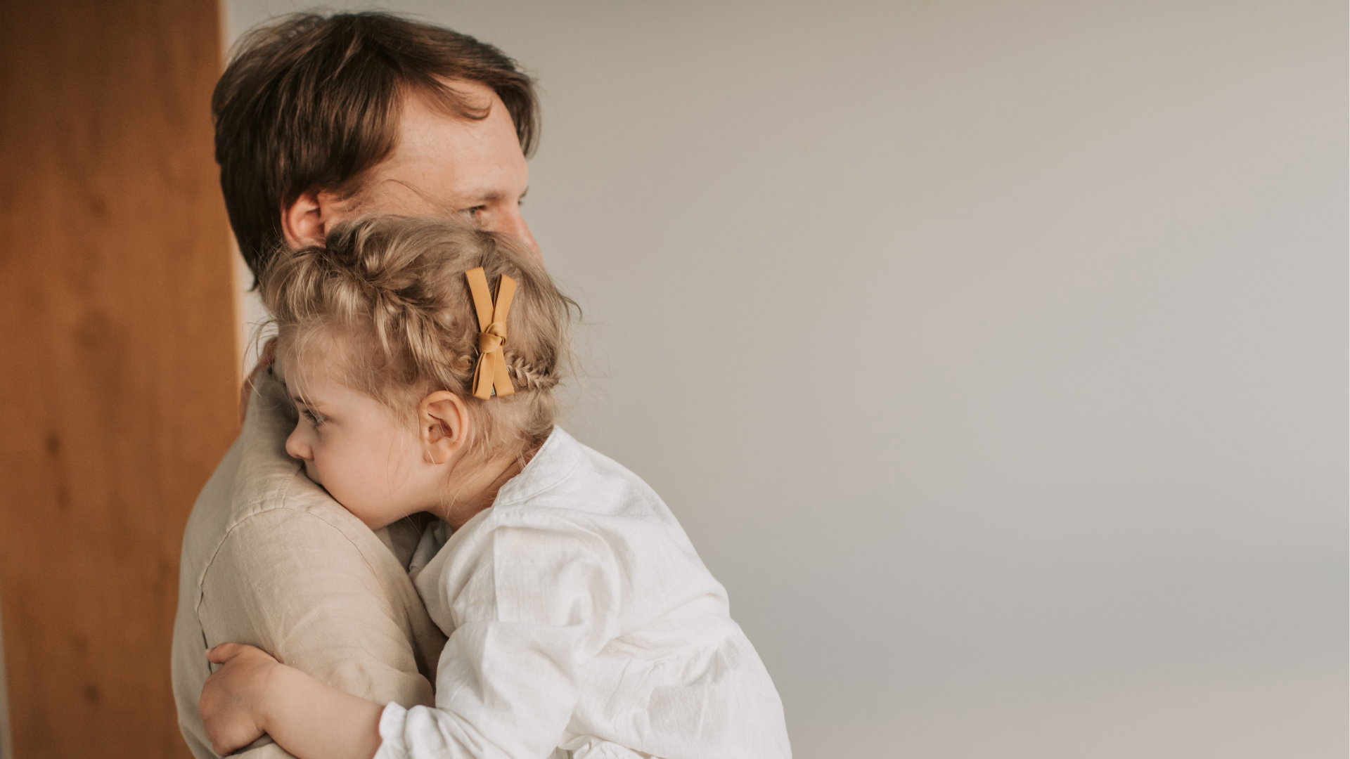 Man wearing tan shirt holding young girl. The girl is wearing a white cotton shirt and has her blonde hair braided with an orange bow tied on the braid.