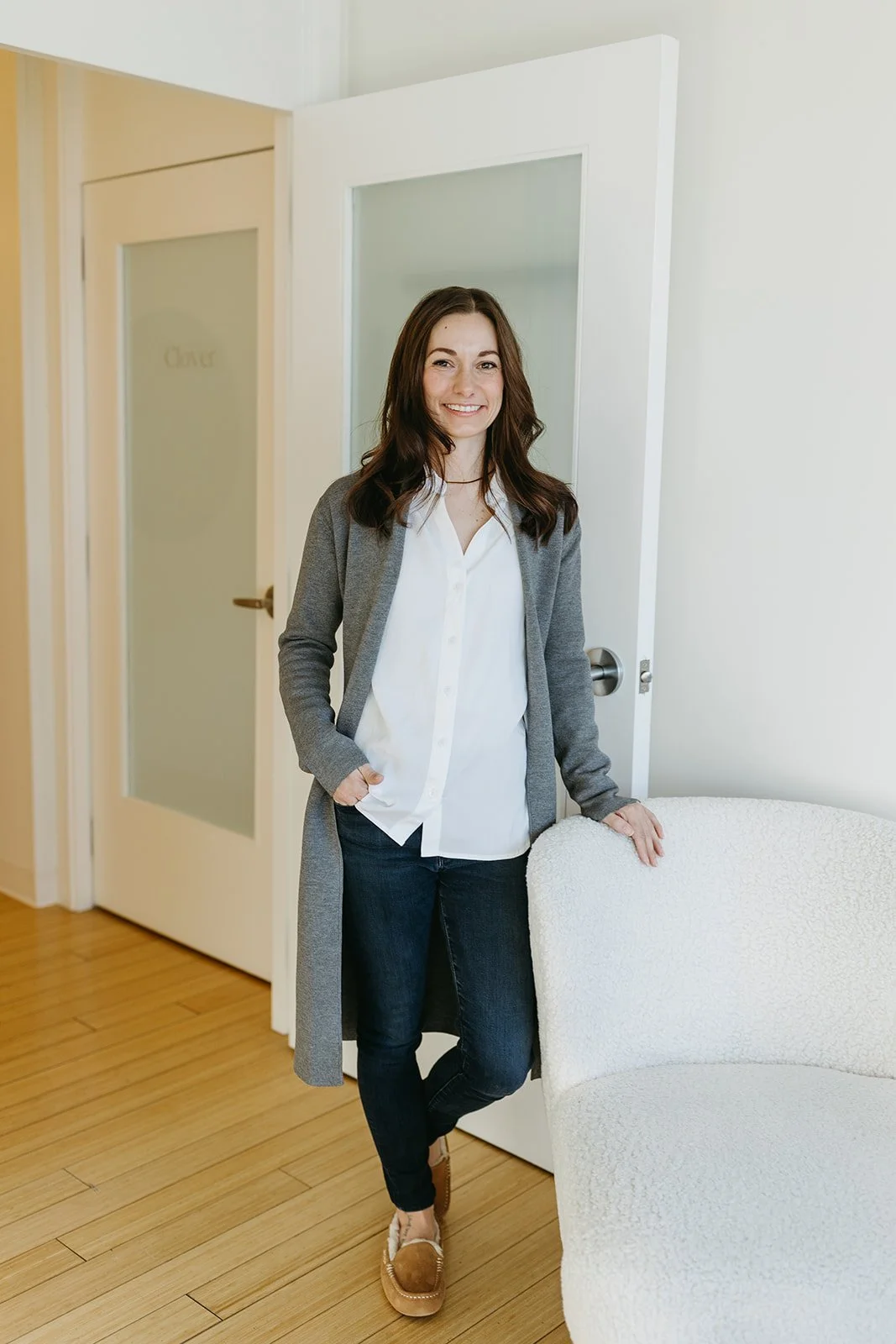 A woman standing indoors, smiling, with dark hair, wearing a gray coat, white blouse, dark jeans, and tan loafers.