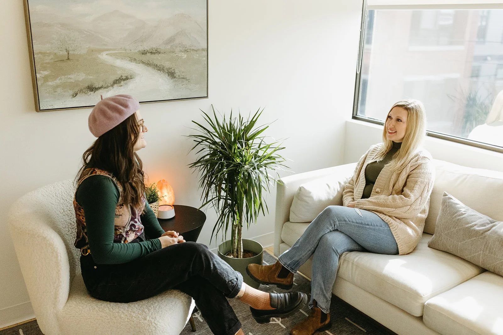 Two women having a conversation in a bright, cozy room with a large window, a potted plant, and abstract artwork on the wall.