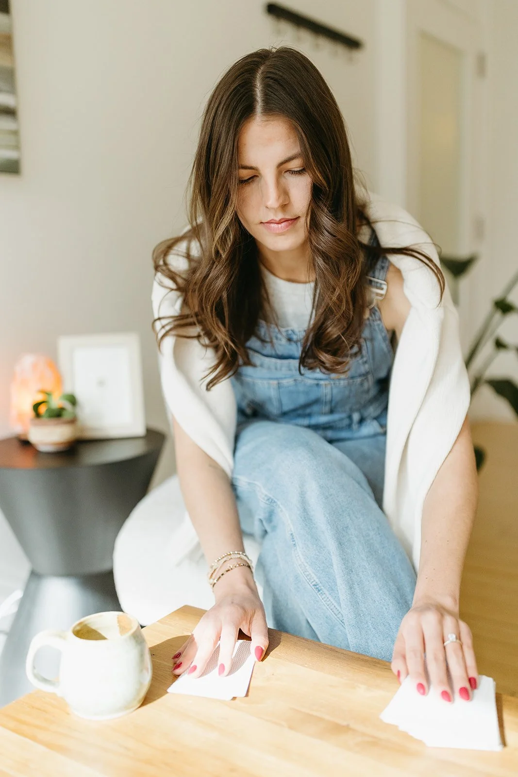 A woman with long brown hair wearing a white shirt and denim overalls, cleaning a wooden table with a cloth in a cozy, well-lit room.