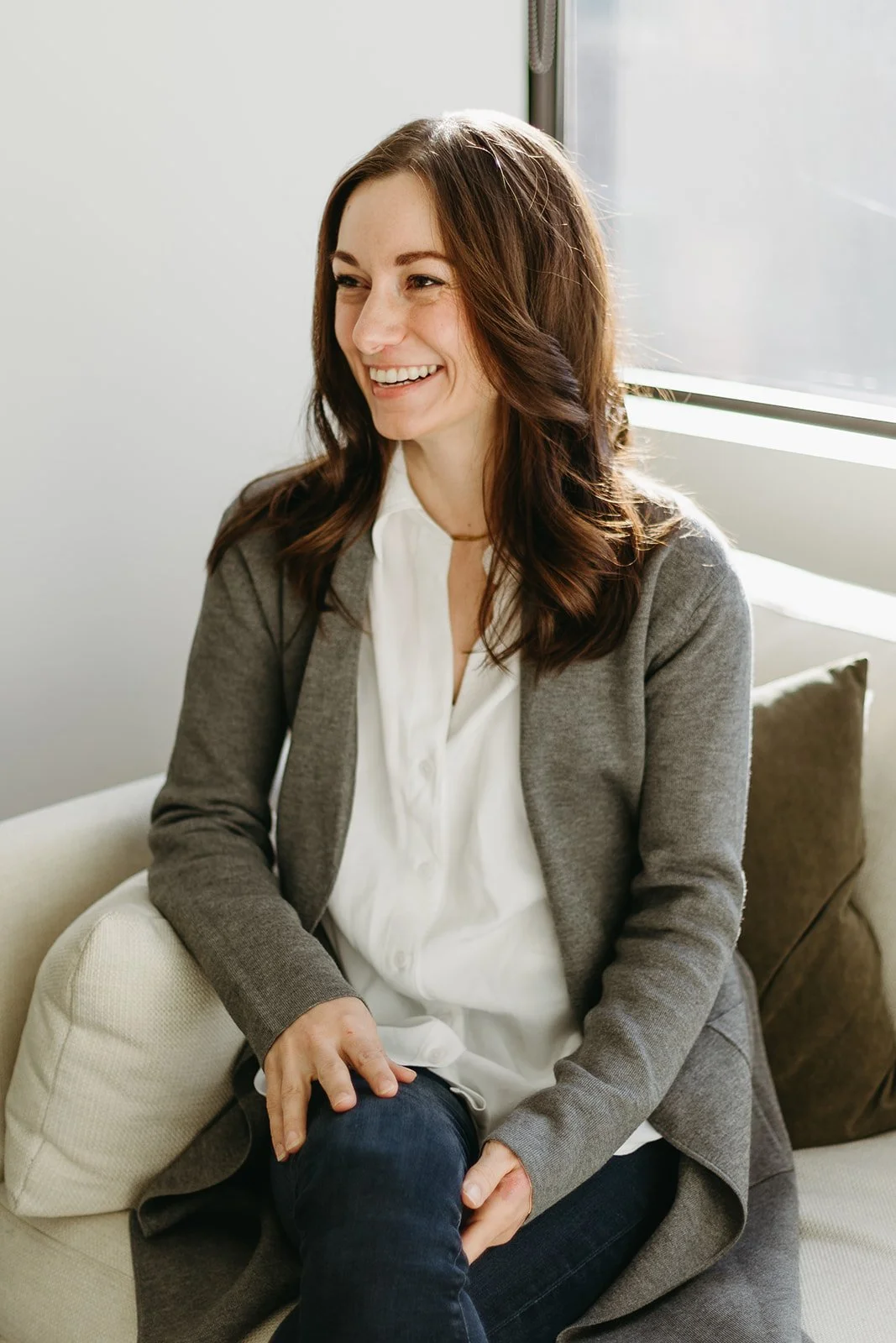 A woman with brown hair smiling, sitting on a sofa by a window, wearing a gray blazer, white blouse, and jeans.