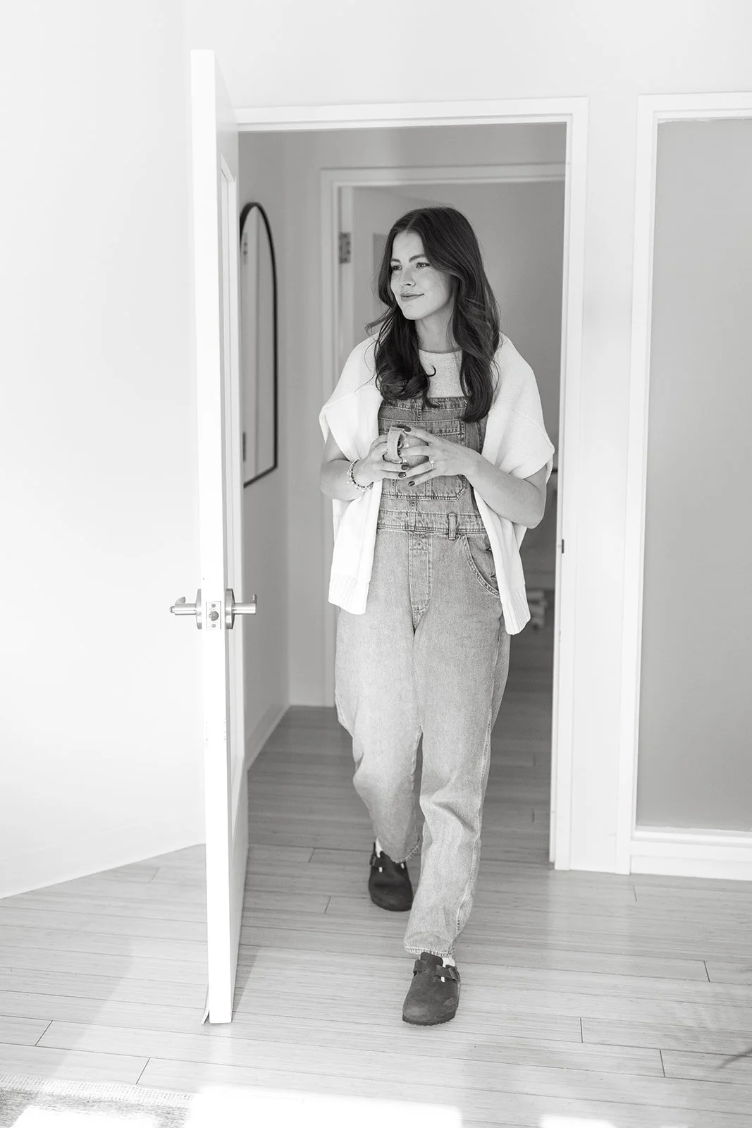 A woman with long wavy hair standing in a doorway, holding a mug, wearing casual overalls and a loose top, in a clean, modern interior.
