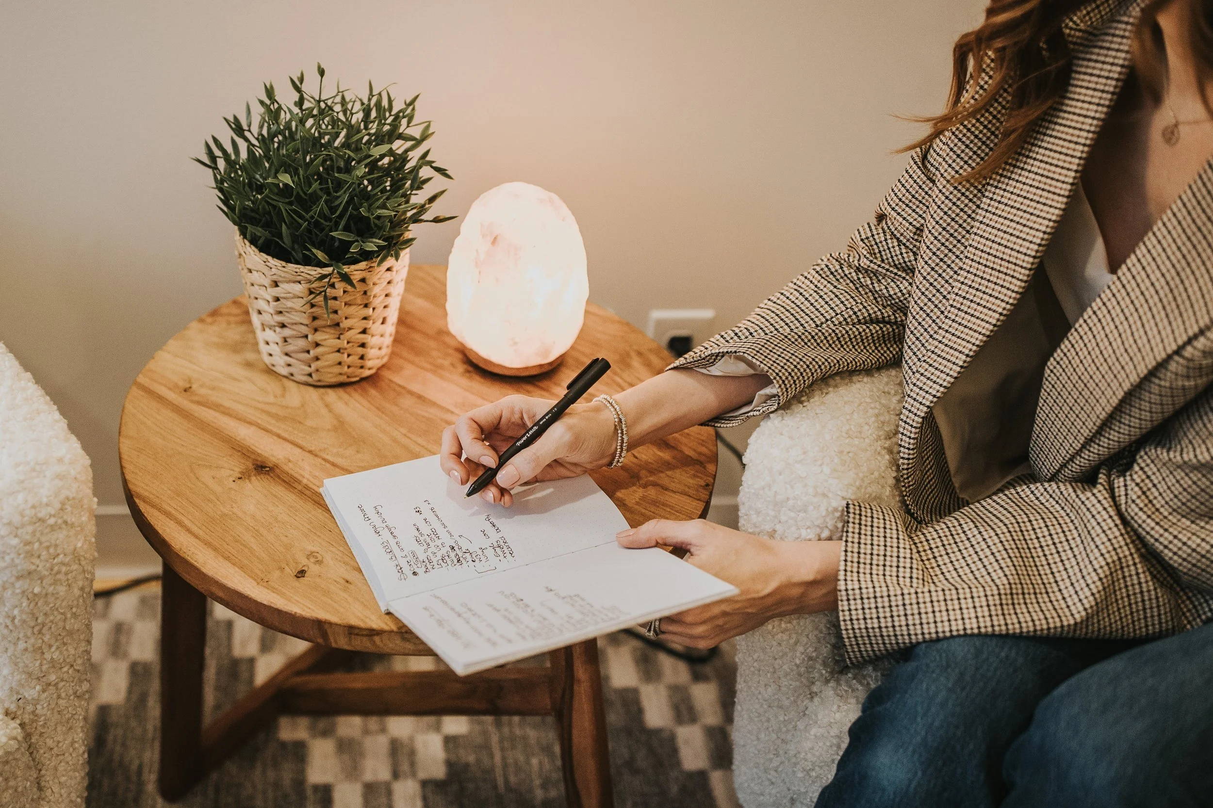 Person sitting on a beige sofa, writing in a notebook on a round wooden table, with a potted plant and a Himalayan salt lamp on the table. Risewell Psychology