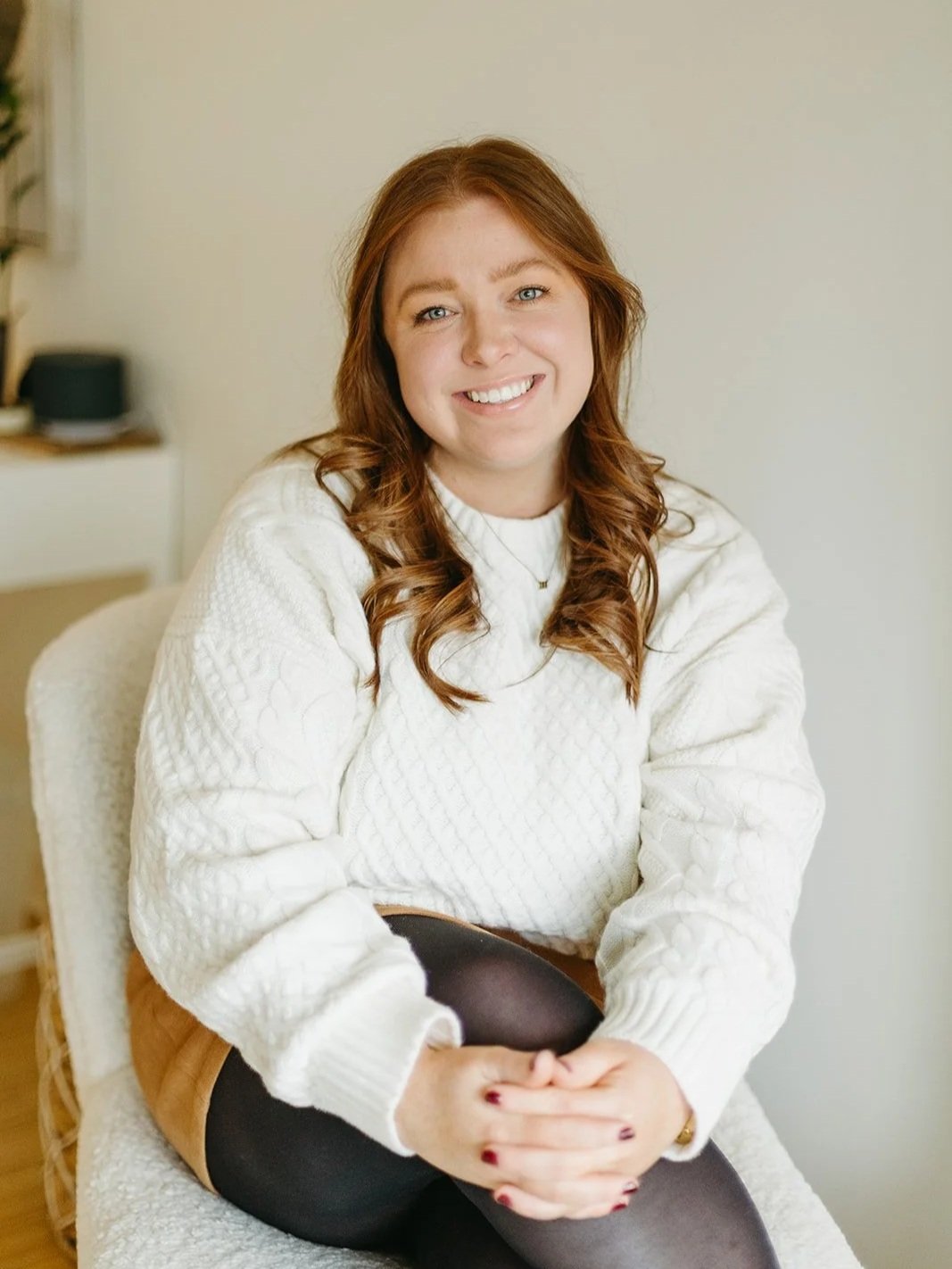 A smiling woman with long curly red hair sitting on a white chair, wearing a white sweater and black tights, in a room with a neutral background.