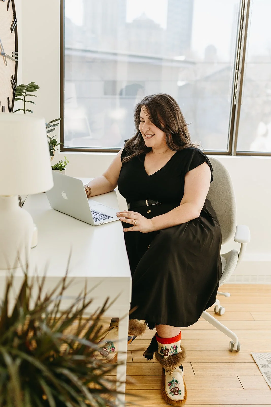 A woman sitting at a desk using a laptop, smiling, wearing black dress and cozy footwear with embroidery, in a bright room with a large window.