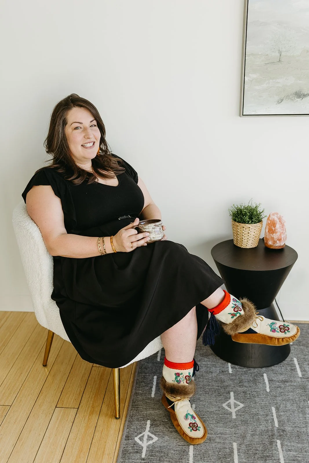 A woman with long brown hair wearing a black dress and colorful socks with moccasin slippers, sitting in a cream-colored chair, holding a mug, and smiling at the camera in a cozy room with modern decor.