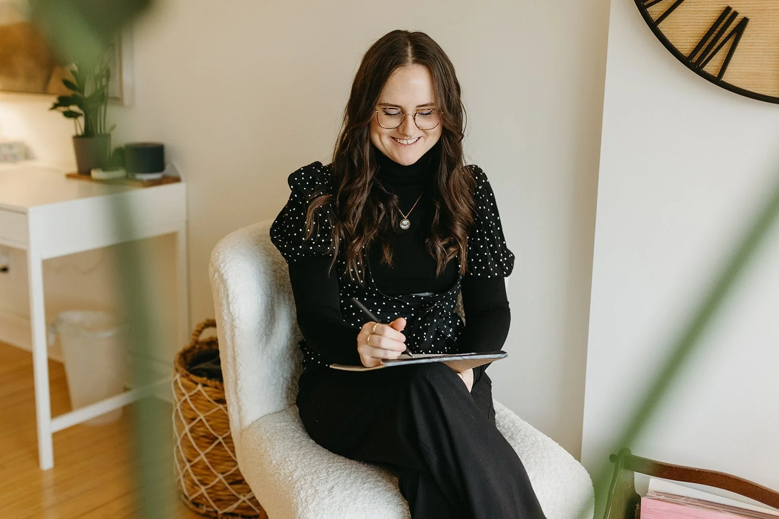 A woman with long brown hair, wearing glasses, a black turtleneck, and a black polka dot dress, sits on a white chair smiling, writing on a small laptop tablet.