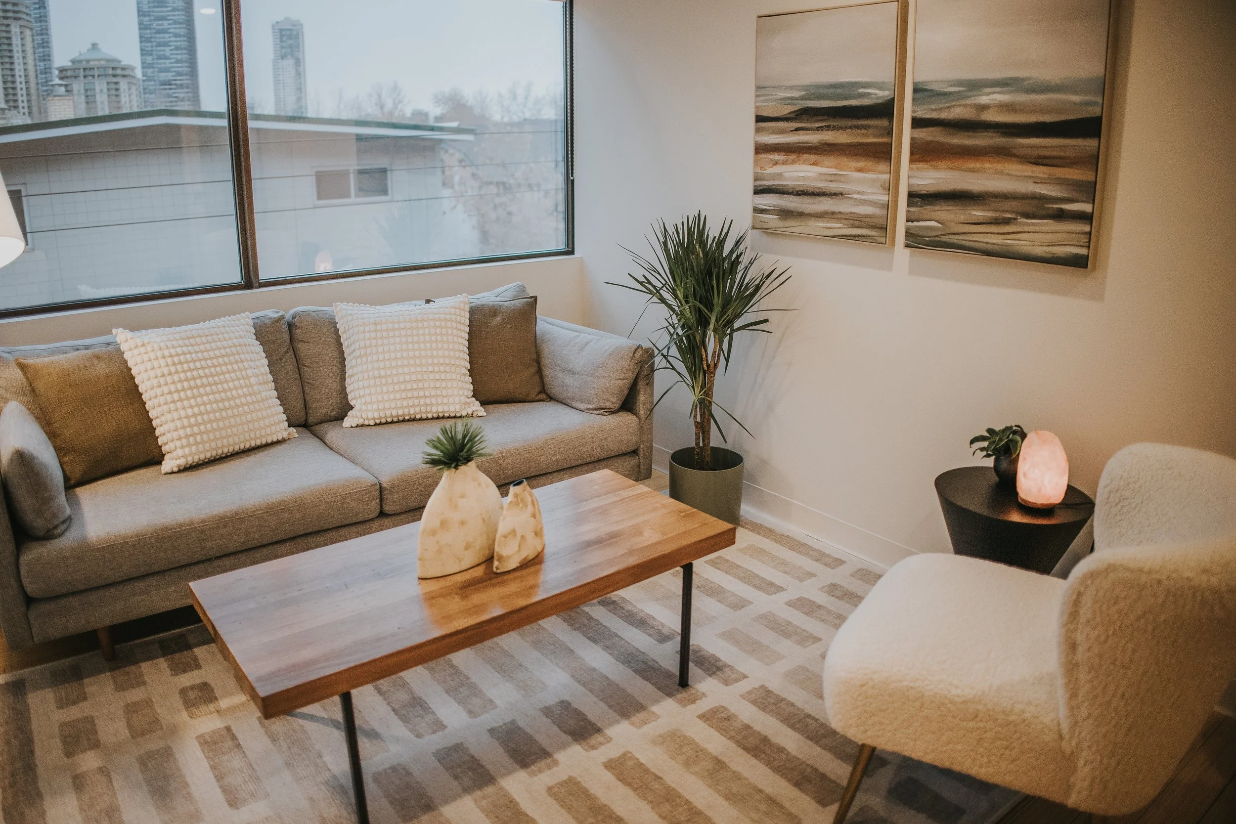 Living room with a gray sofa, white textured pillows, a wooden coffee table with decorative stones and plants, a potted plant in a corner, a beige armchair, a side table with a pink salt lamp, abstract landscape paintings on the wall, and a large window with a city view.