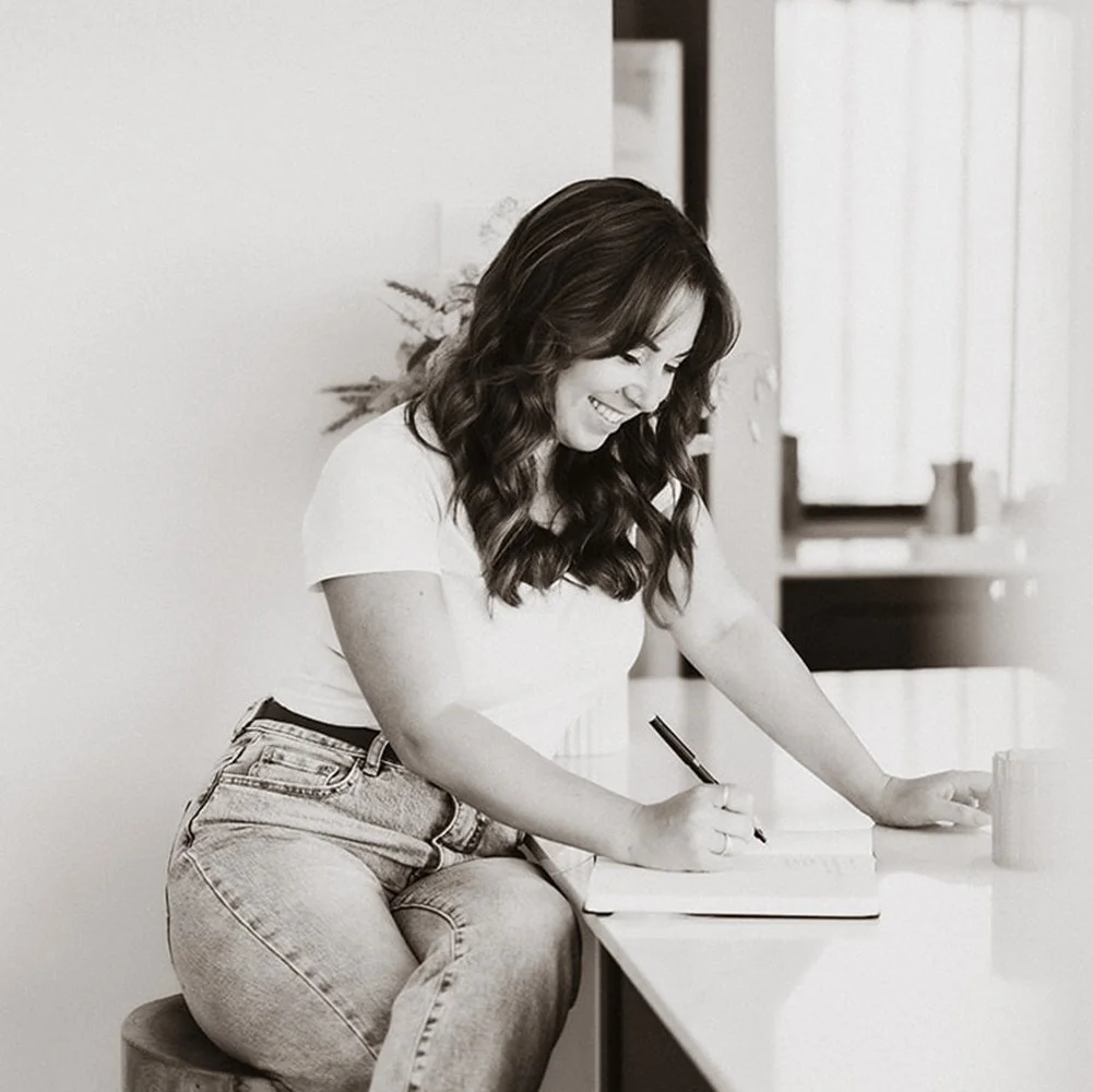 Black and white image of graphic designer sitting at kitchen bench taking notes.