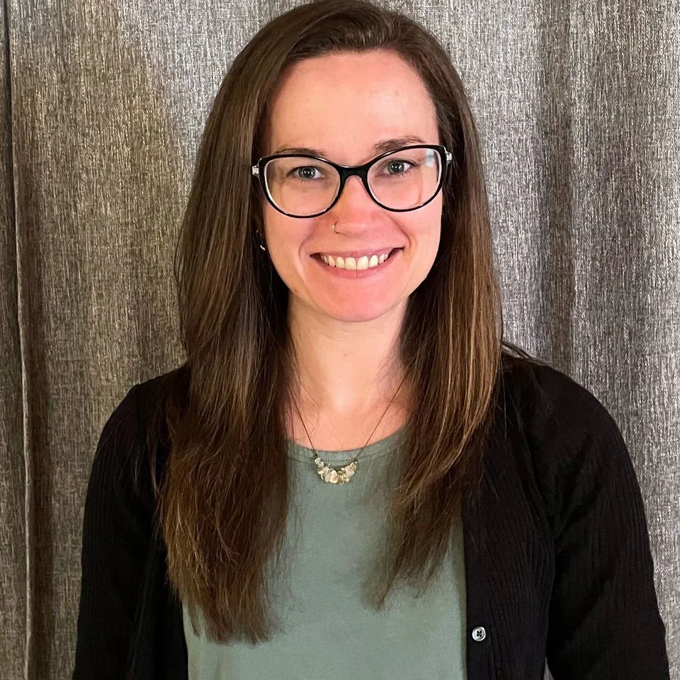 A smiling woman with long brown hair, wearing glasses and a nose piercing, in front of a textured gray background.