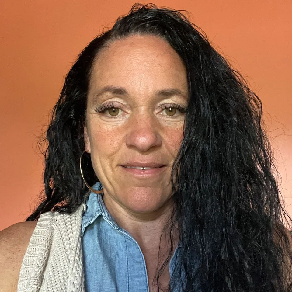 A woman with long, curly black hair wearing a sleeveless, textured knit sweater over a blue collared shirt, standing against an orange background.