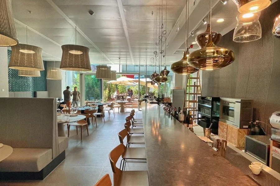 Modern restaurant interior with bar and dining tables, large pendant lights, and outdoor seating area visible through glass windows.