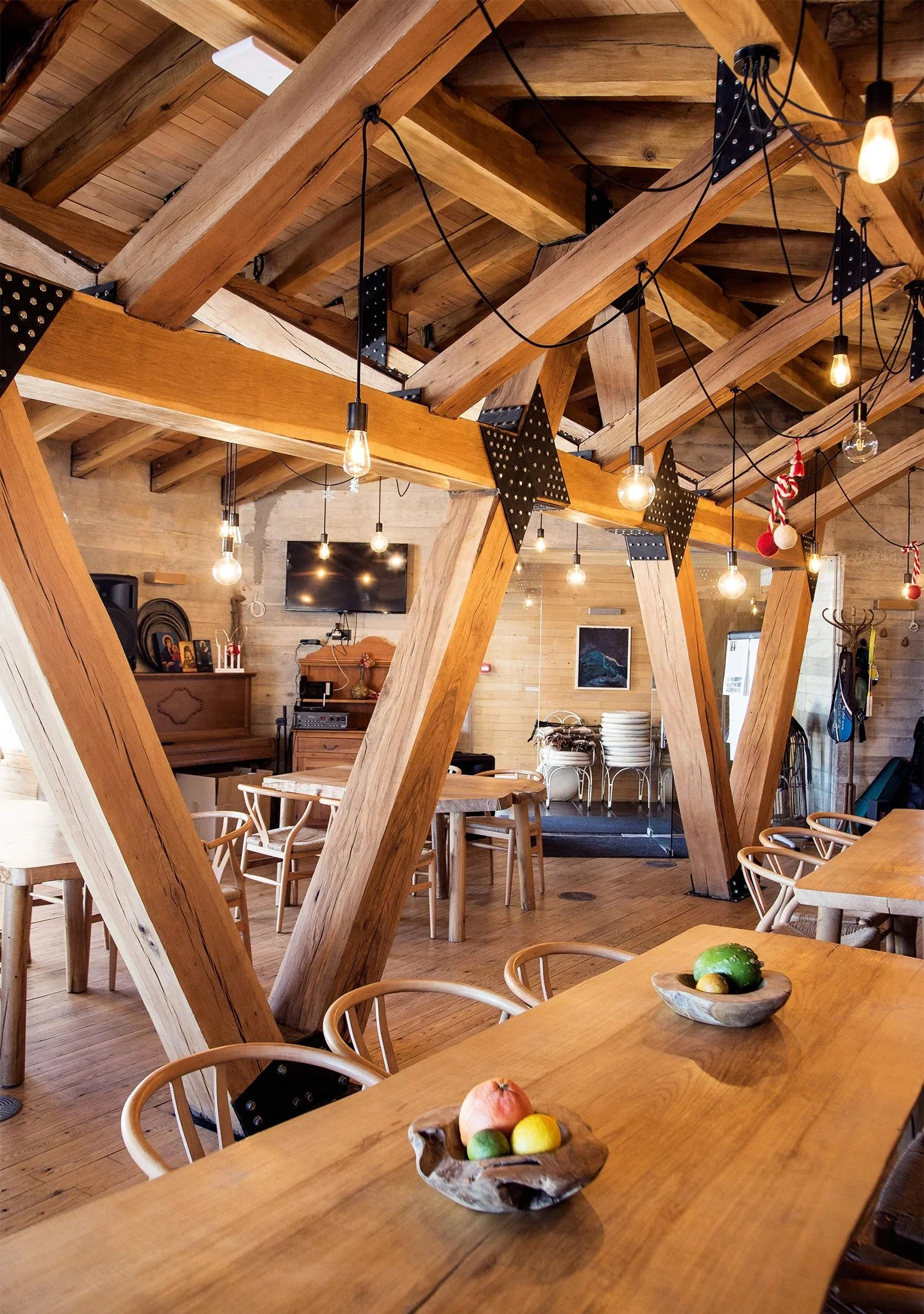 Rustic interior of a restaurant with wooden beams, tables, and chairs, featuring hanging light bulbs and decorative fruit bowls on tables.