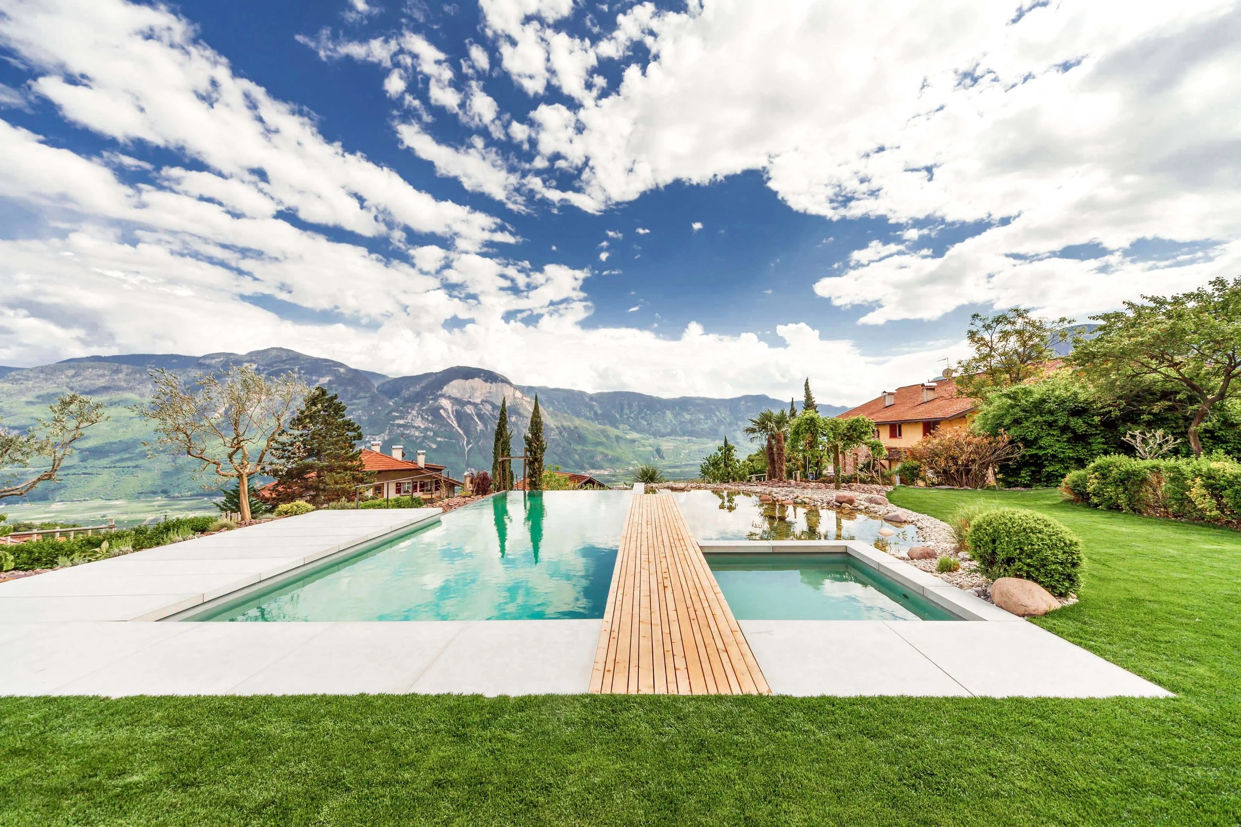 Ein Pool mit Blick auf Berge, umgeben von grüner Rasenfläche und Büschen, mit einem Holzsteg in der Mitte.