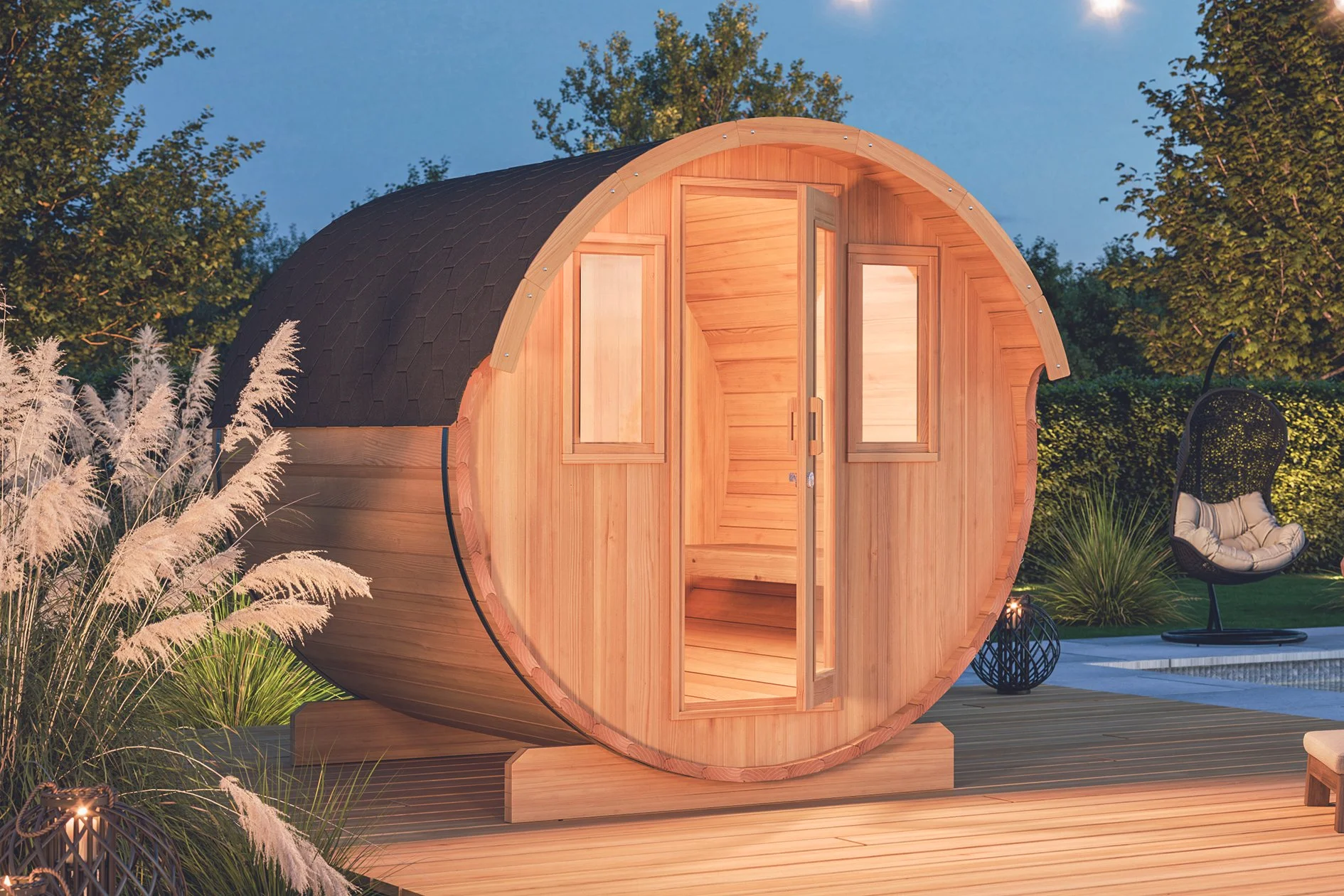 Ein kleines, runden Holzhaus mit einer halboffenen Tür, auf einer Holzterrasse, umgeben von Pflanzen und einer Gartenliege bei Dämmerung.