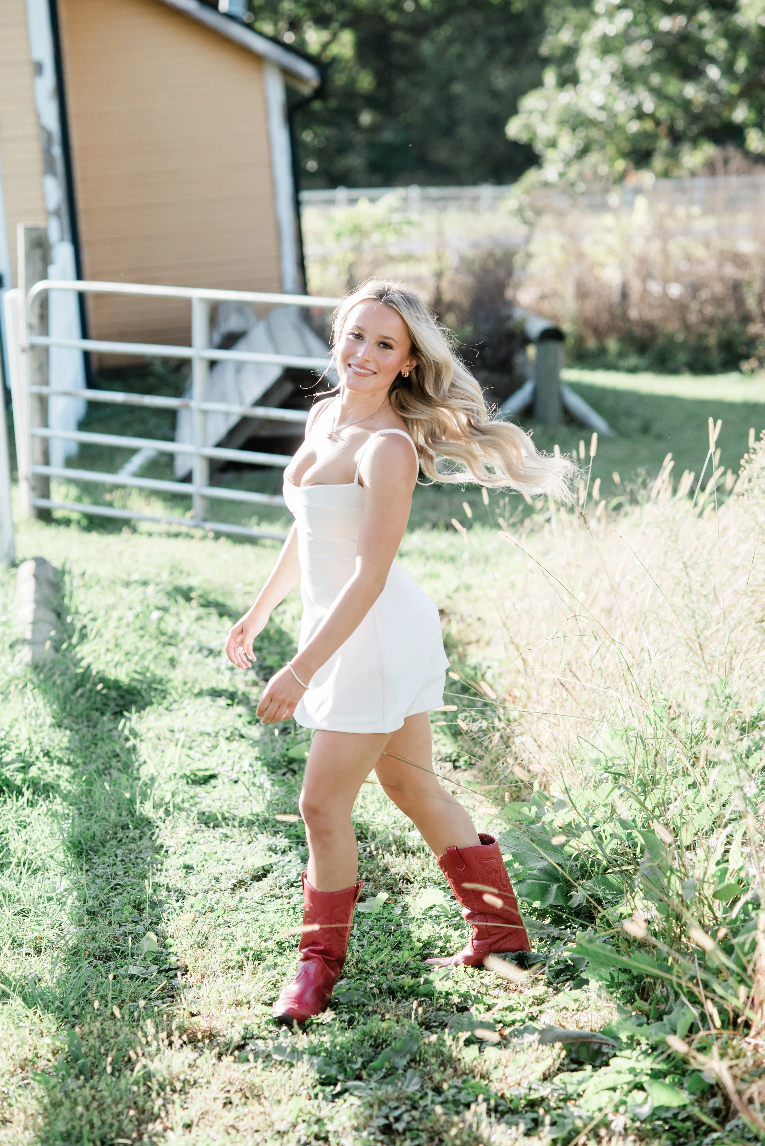 A young woman with long blonde hair wearing a white sleeveless dress and red cowboy boots standing outdoors in a grassy area with trees and a brown building in the background.