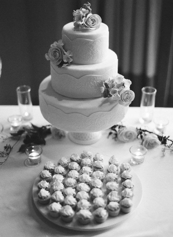 A three-tiered wedding cake decorated with roses on top, with a plate of cupcakes in front, surrounded by candles on a table.