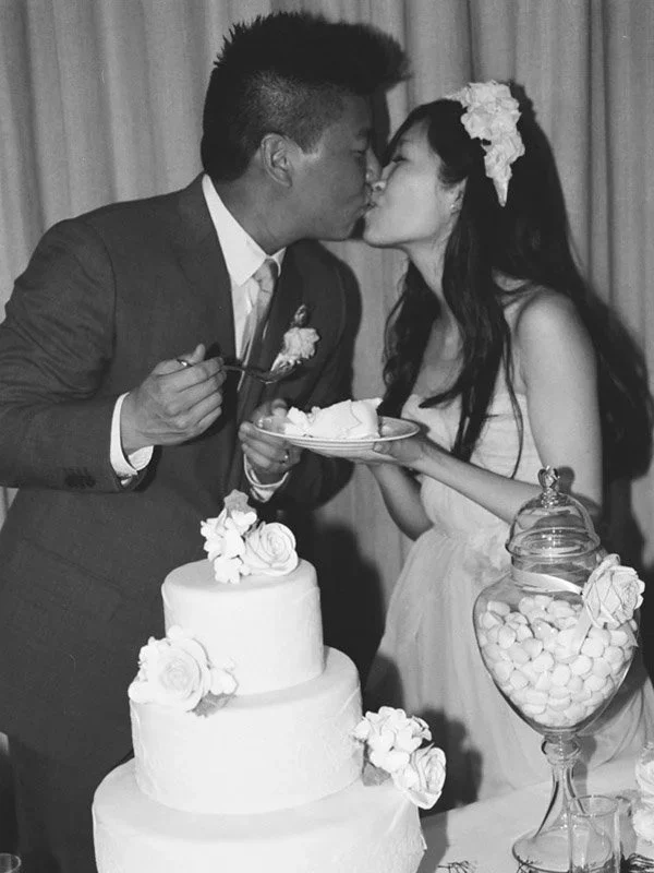 A black and white photo of a couple at their wedding, kissing over a wedding cake, with a table of candy in a jar in front of them.