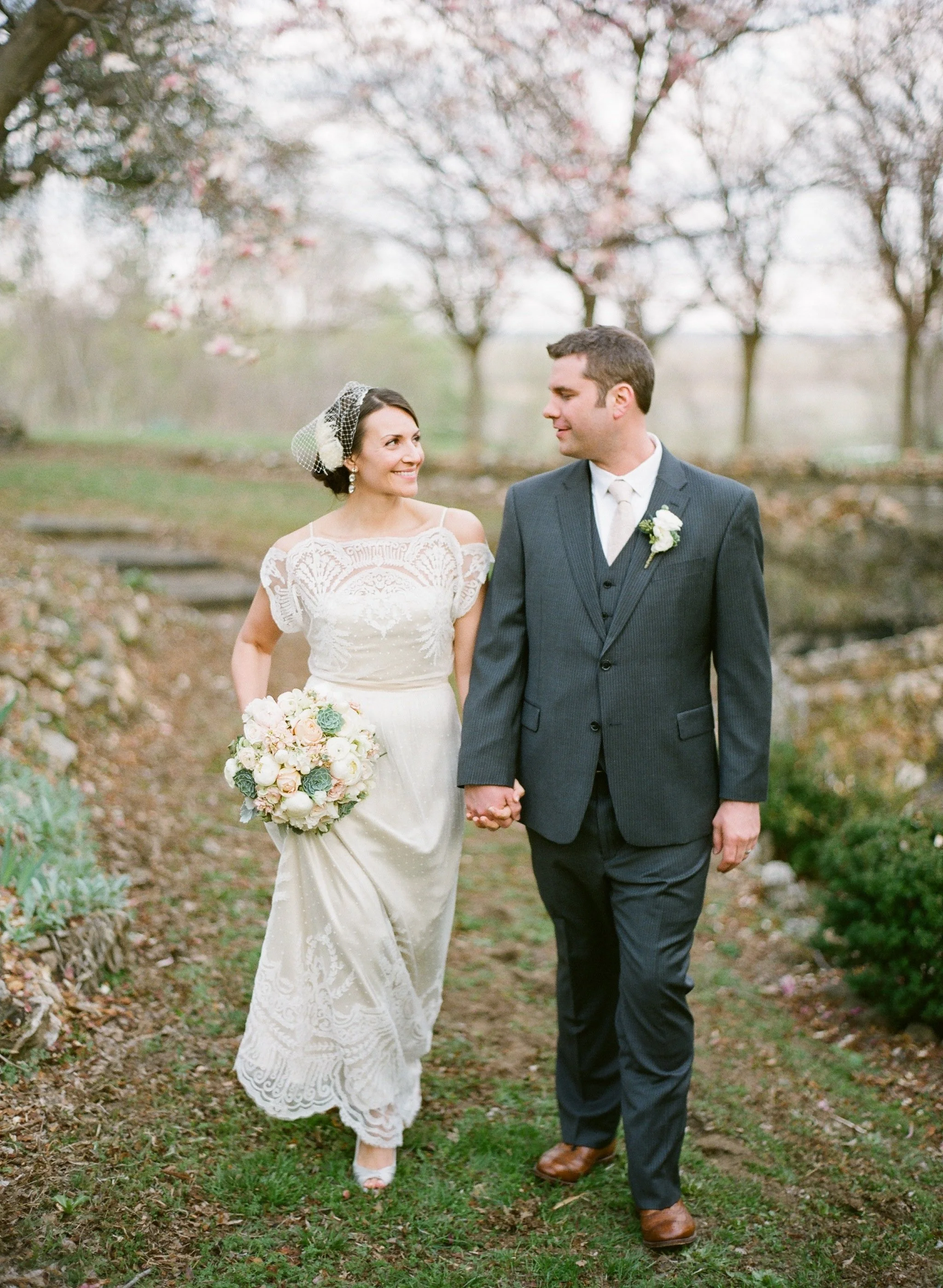 A bride and groom walking outdoors on a garden path, holding hands, surrounded by blooming trees, during a wedding celebration.