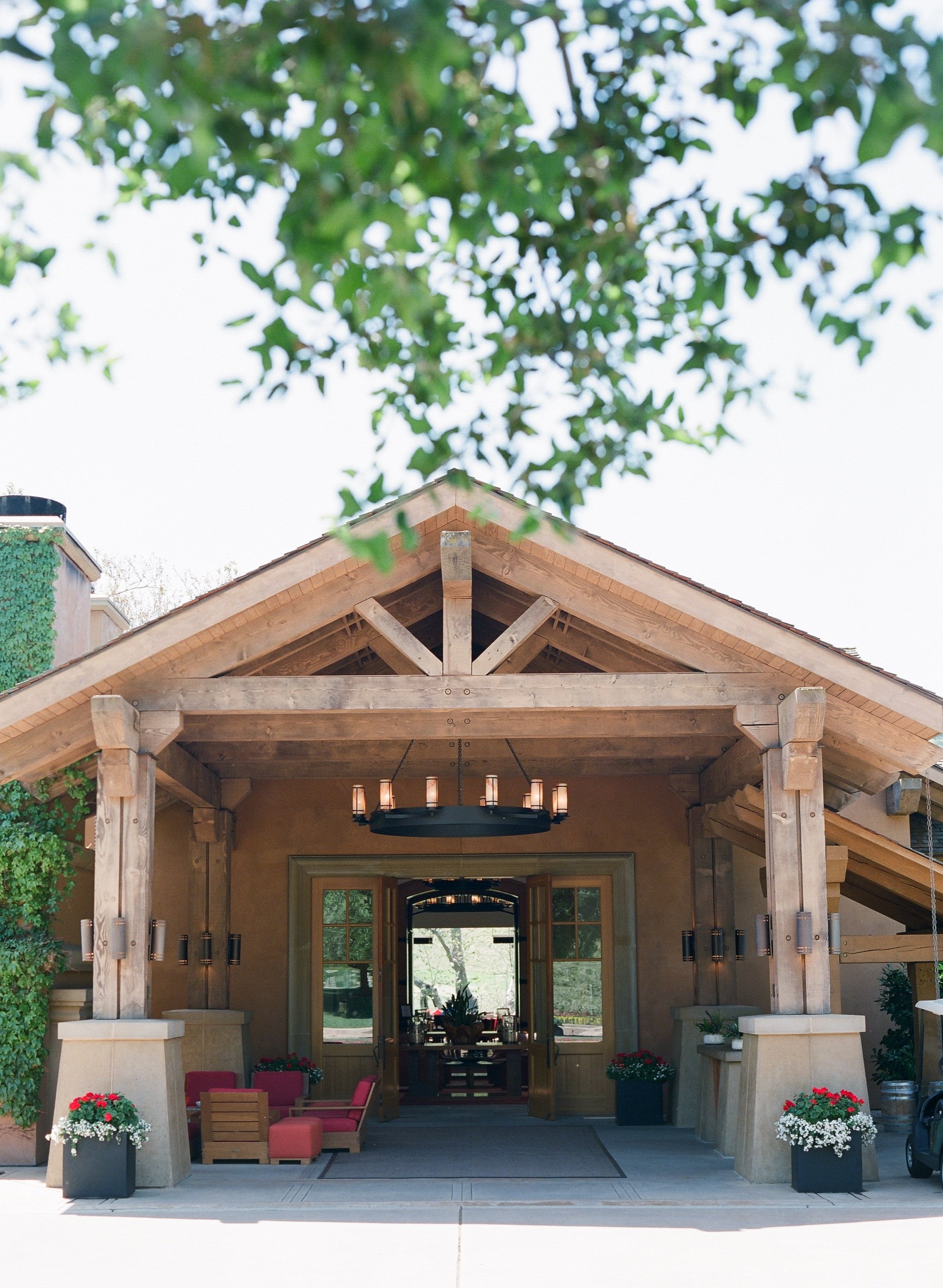 Front entrance of a house with a large wooden porch, hanging chandelier, red chairs, and potted flowers, framed by green trees.