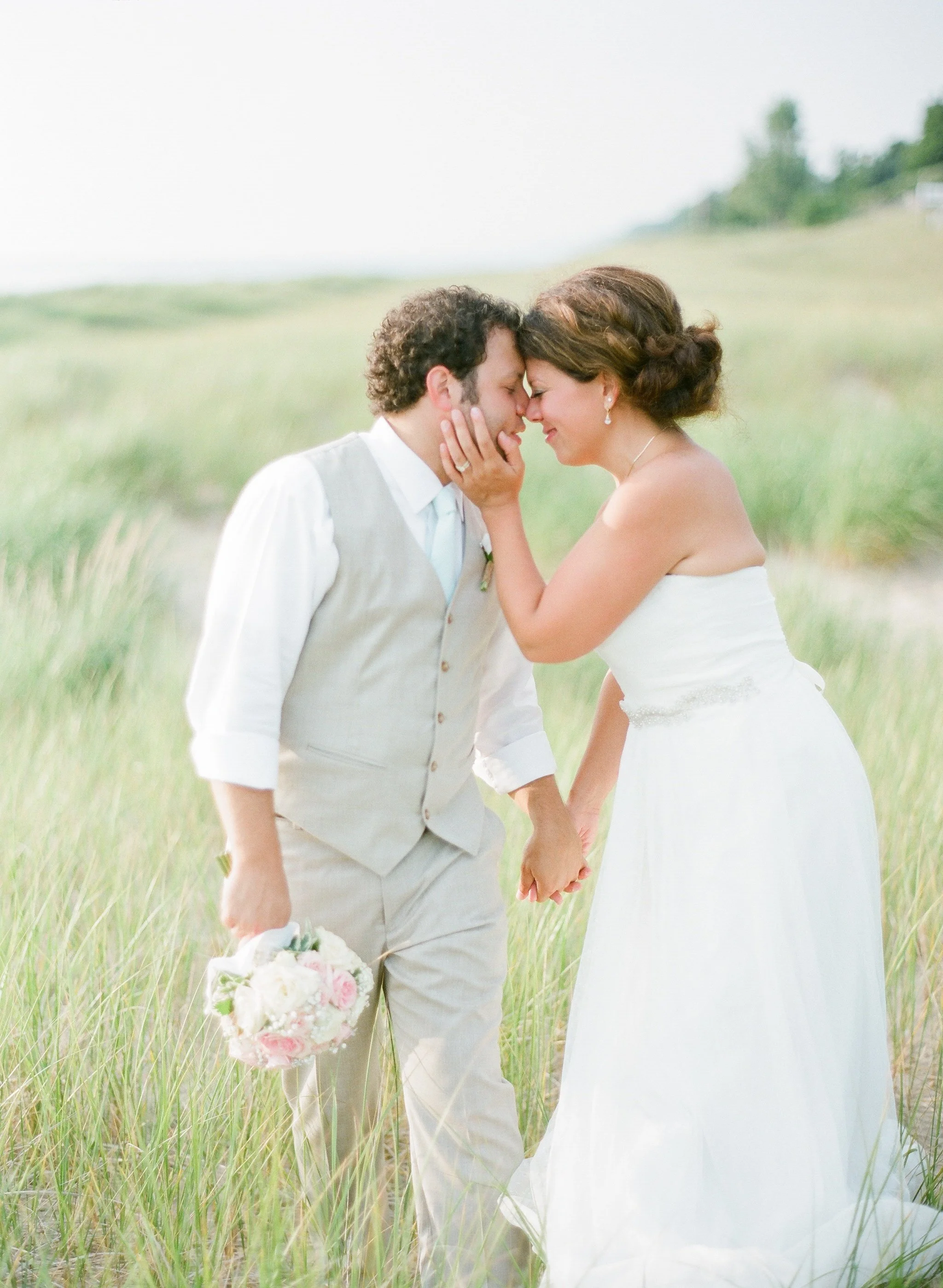 A bride and groom holding hands and touching foreheads in a grassy field, dressed in wedding attire, romantic moment.