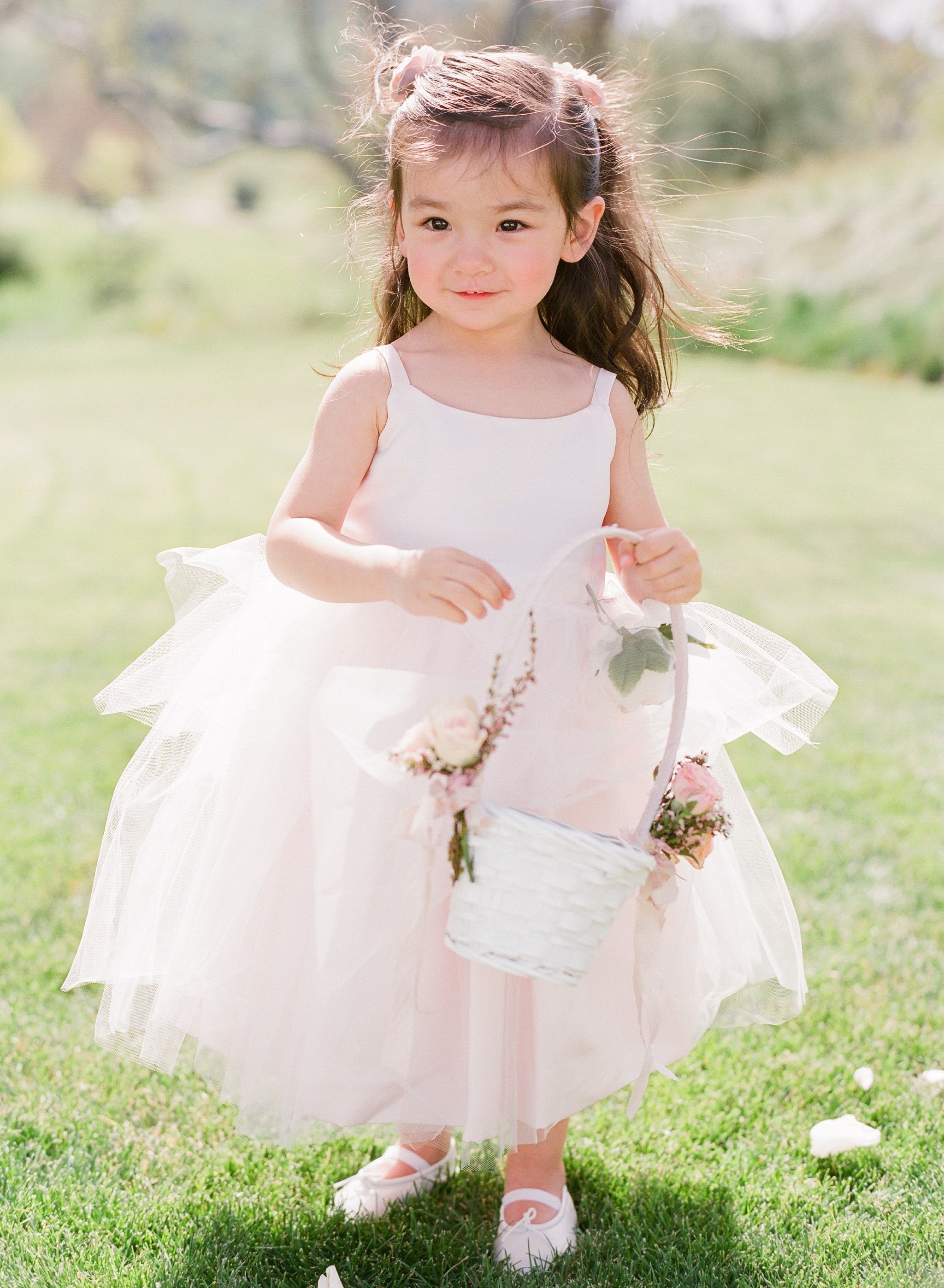 A young girl in a white dress with a tulle skirt, holding a white basket with pink flowers, standing on grass outdoors on a sunny day