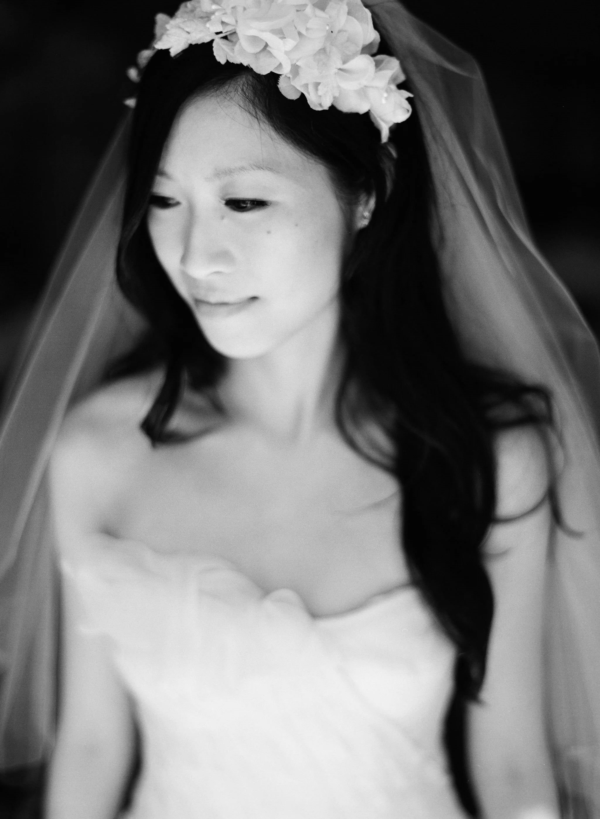 Black and white photo of a woman with flowers in her hair, wearing a veil, and looking down.