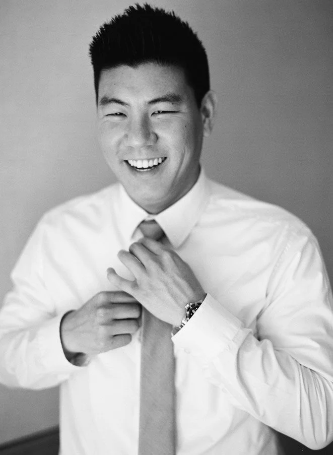 A smiling Asian man in a white dress shirt and tie adjusting his tie, wearing a watch, in a professional setting.