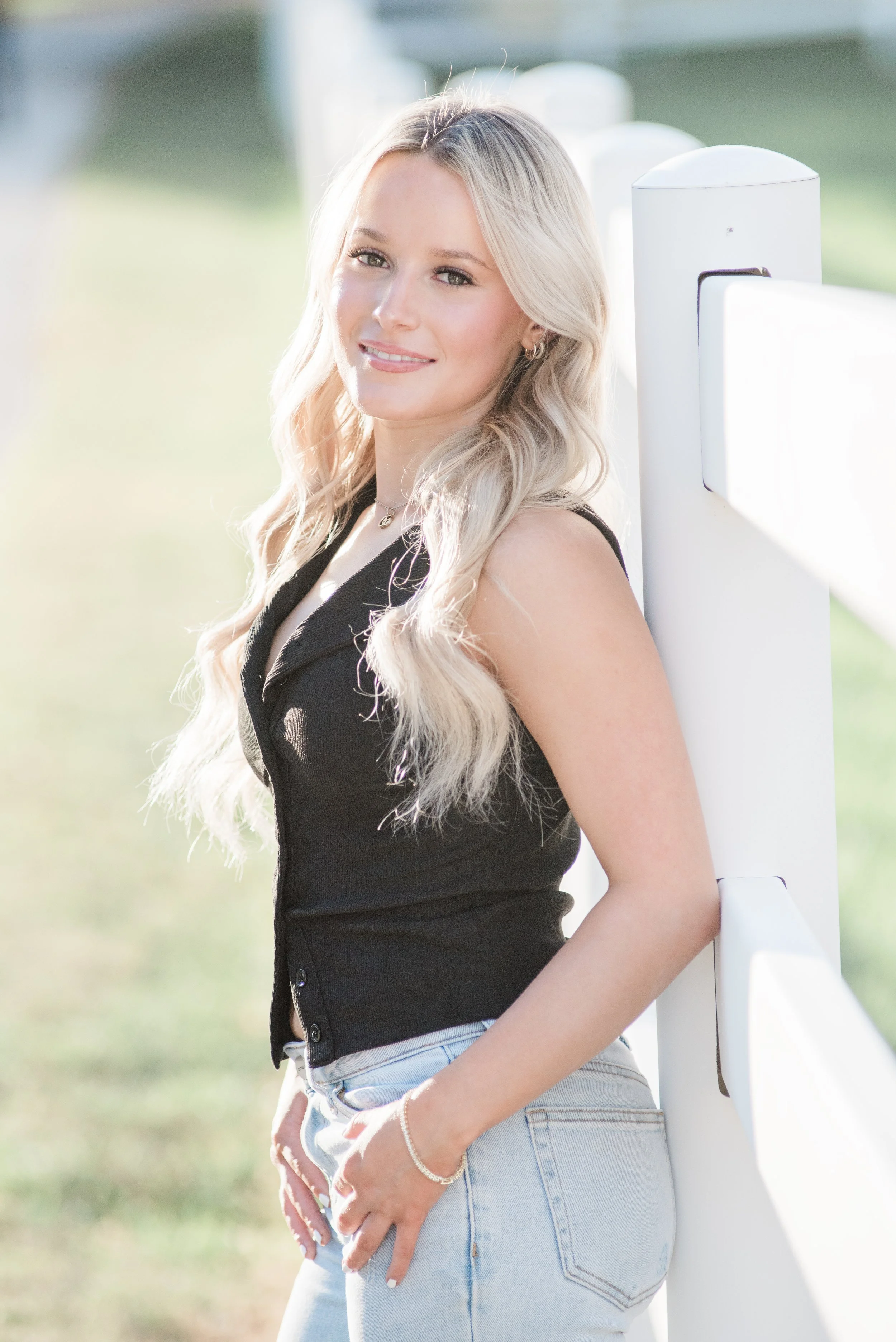 Young woman with long blond hair standing outdoors beside a white fence, wearing a black sleeveless top and light denim jeans.