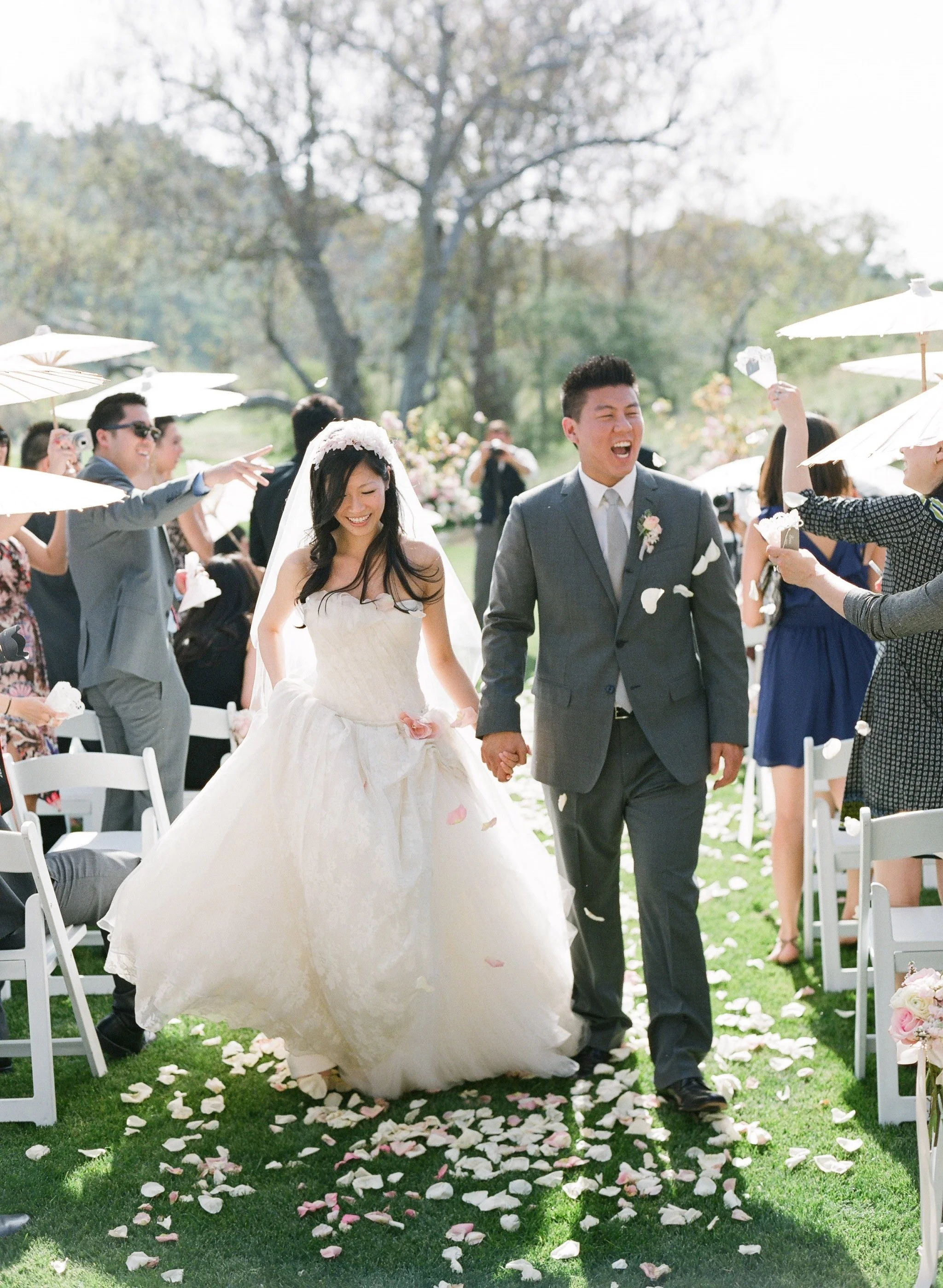 A bride and groom holding hands, walking down an outdoor aisle at their wedding ceremony, surrounded by guests under umbrellas, with flower petals scattered on the grass.