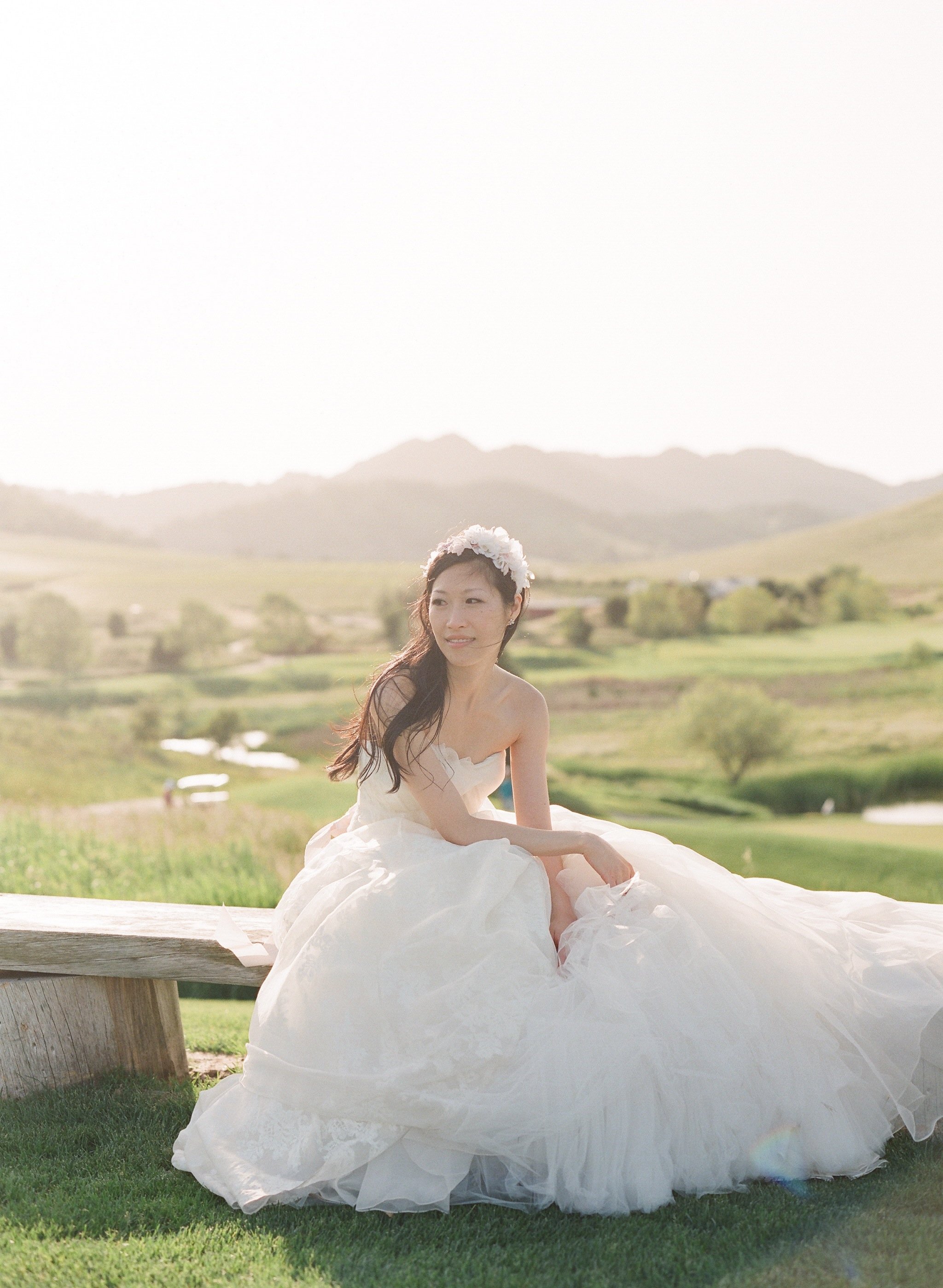 A woman in a wedding dress sitting on a wooden bench outdoors with a landscape of hills and trees in the background, during daytime.