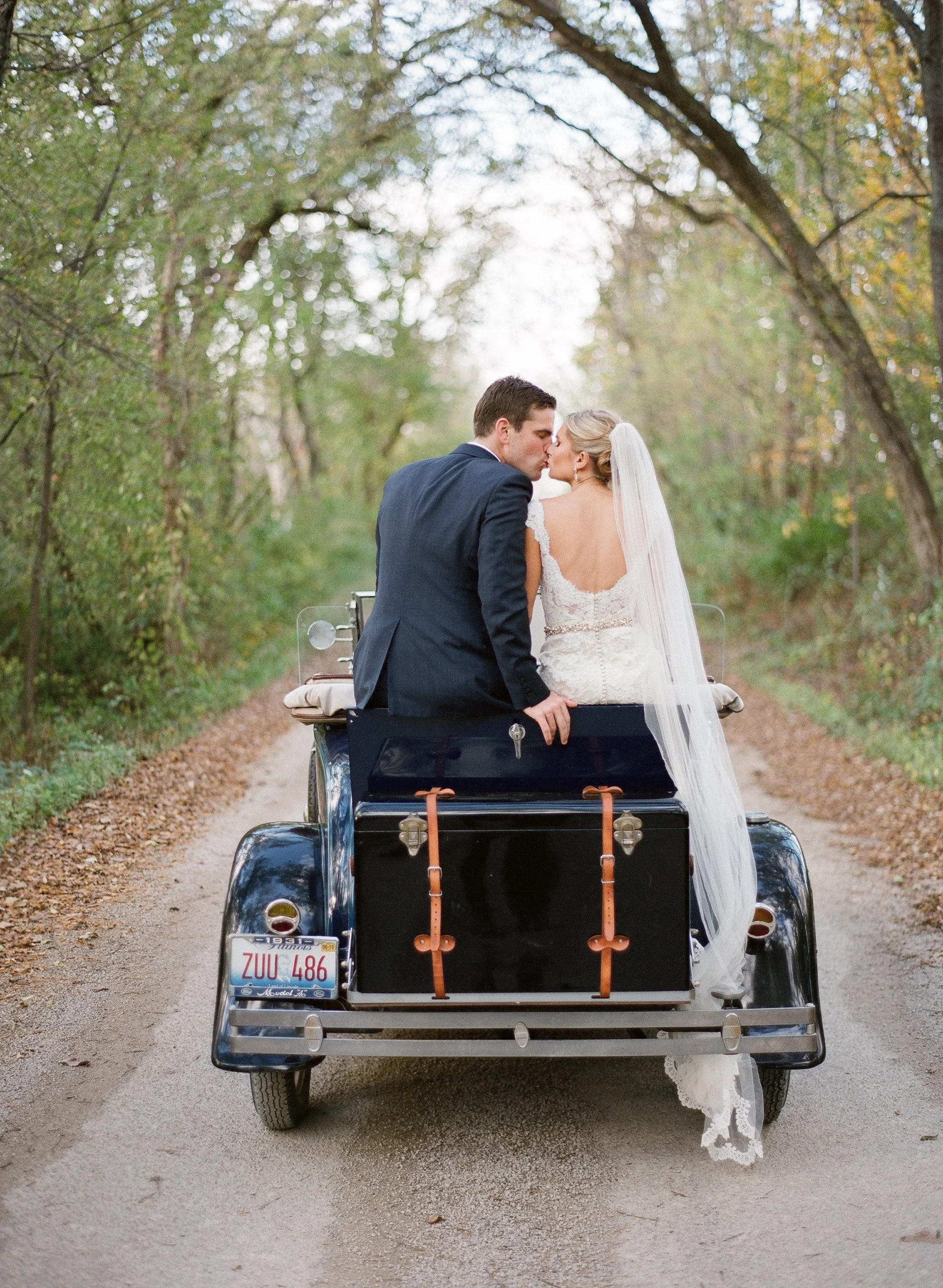 A bride and groom kissing while riding in a vintage car on a dirt road surrounded by trees with fall foliage.