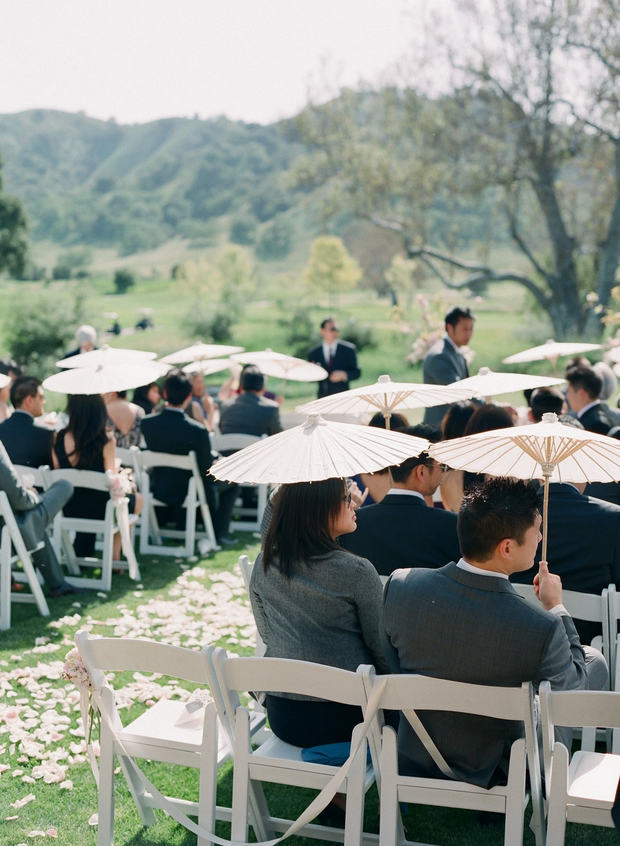 People attending an outdoor wedding ceremony sitting on white chairs with white umbrellas providing shade, in a lush green garden with trees and hills in the background.