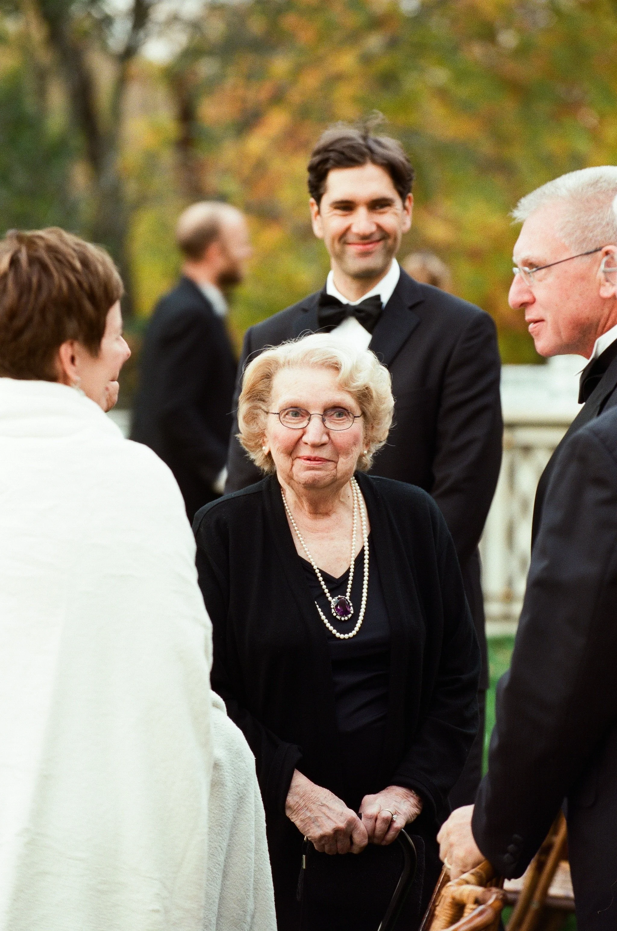A woman wearing glasses and a black dress with a pearl necklace, talking to a man in a tuxedo, with other people in the background at an outdoor event during autumn.