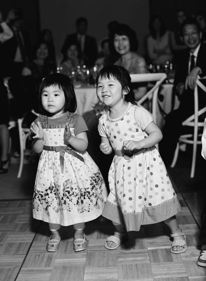 Two young girls in floral dresses dancing during a social gathering with people seated at tables in the background.