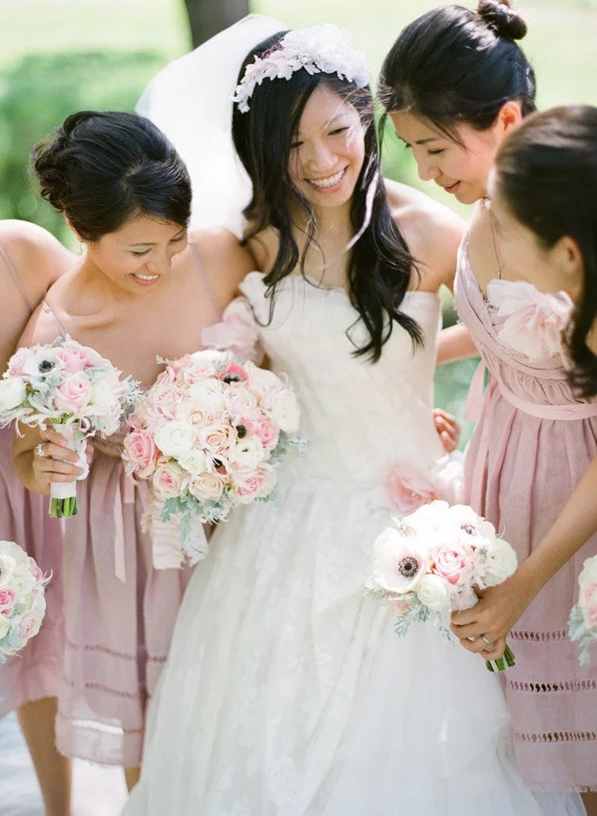 Smiling bride in a white wedding dress surrounded by bridesmaids in pink dresses, holding bouquets of pink and white roses, outdoors in a park setting.