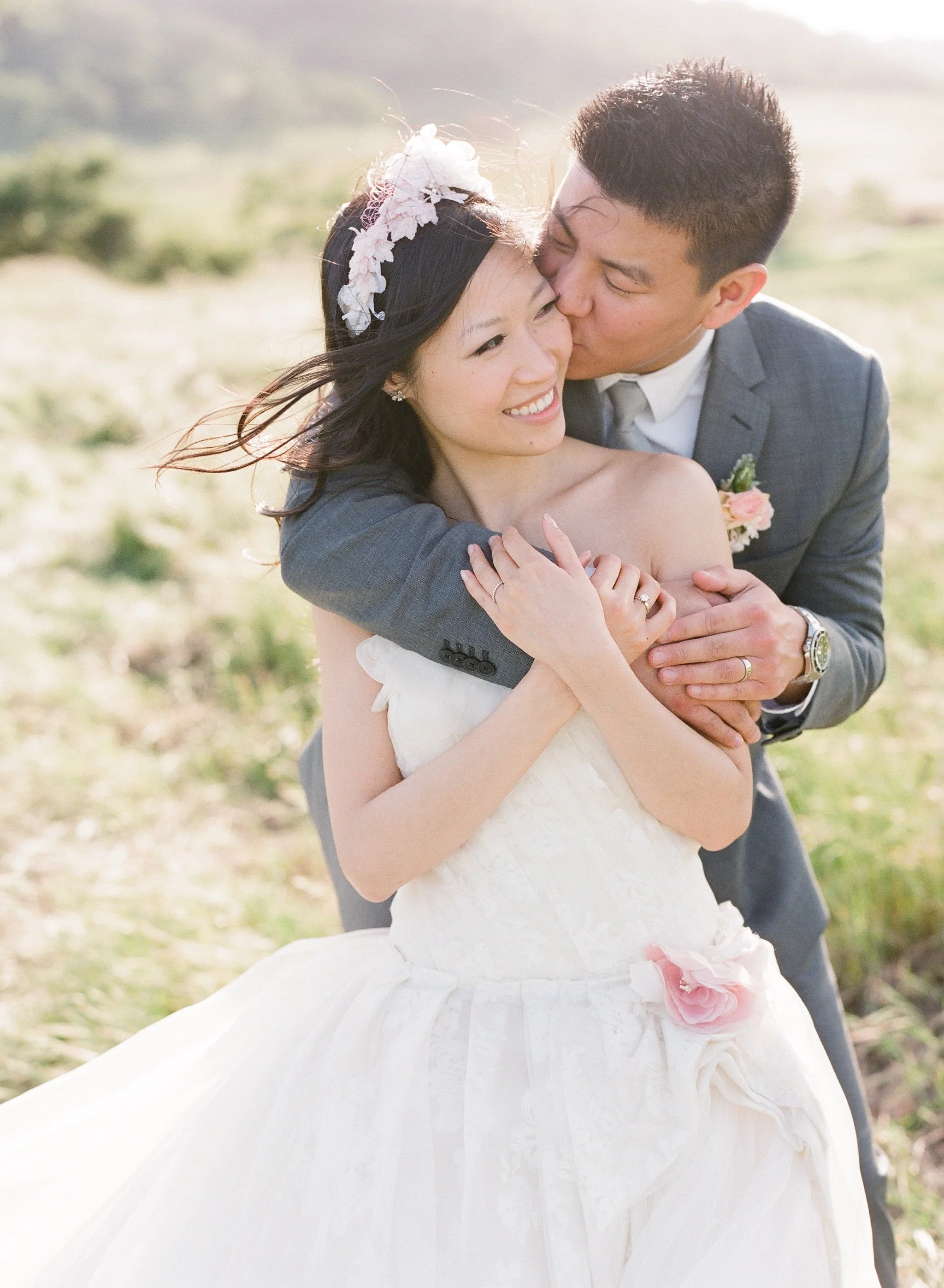 A bride and groom embracing outdoors, with the groom kissing the bride on the cheek. The bride is smiling and wearing a white wedding dress with a floral headpiece, while the groom is dressed in a gray suit with a boutonniere.