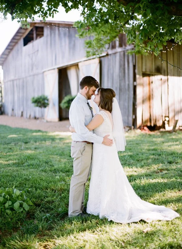 Bride and groom embrace outdoors in front of a rustic barn on their wedding day, under a tree with string lights.