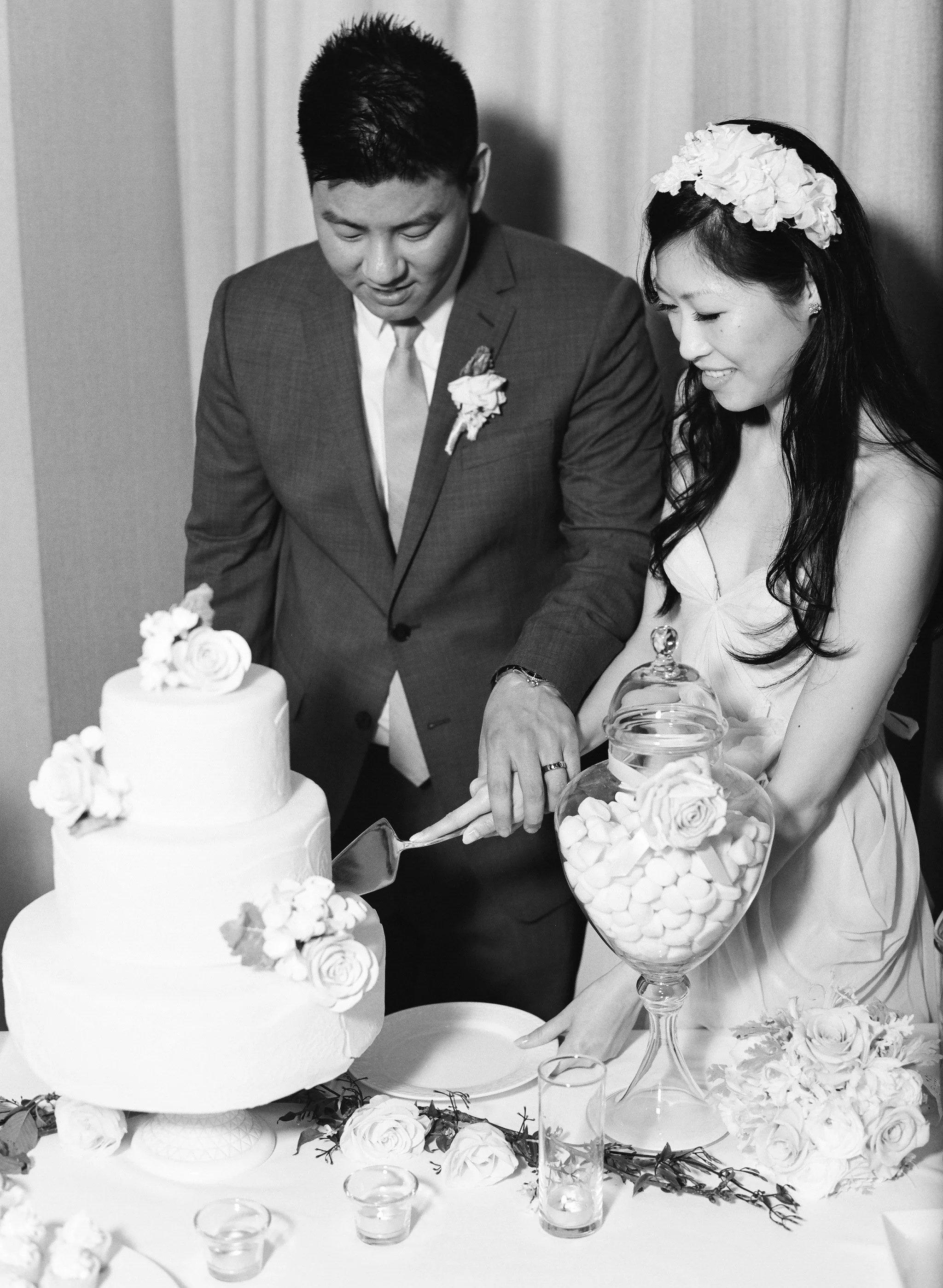 A black-and-white photo of a bride and groom cutting a wedding cake, decorated with flowers. The bride is wearing a dress and a floral headpiece, and the groom is in a suit with a boutonniere. They are smiling and focused on the cake as they cut it t