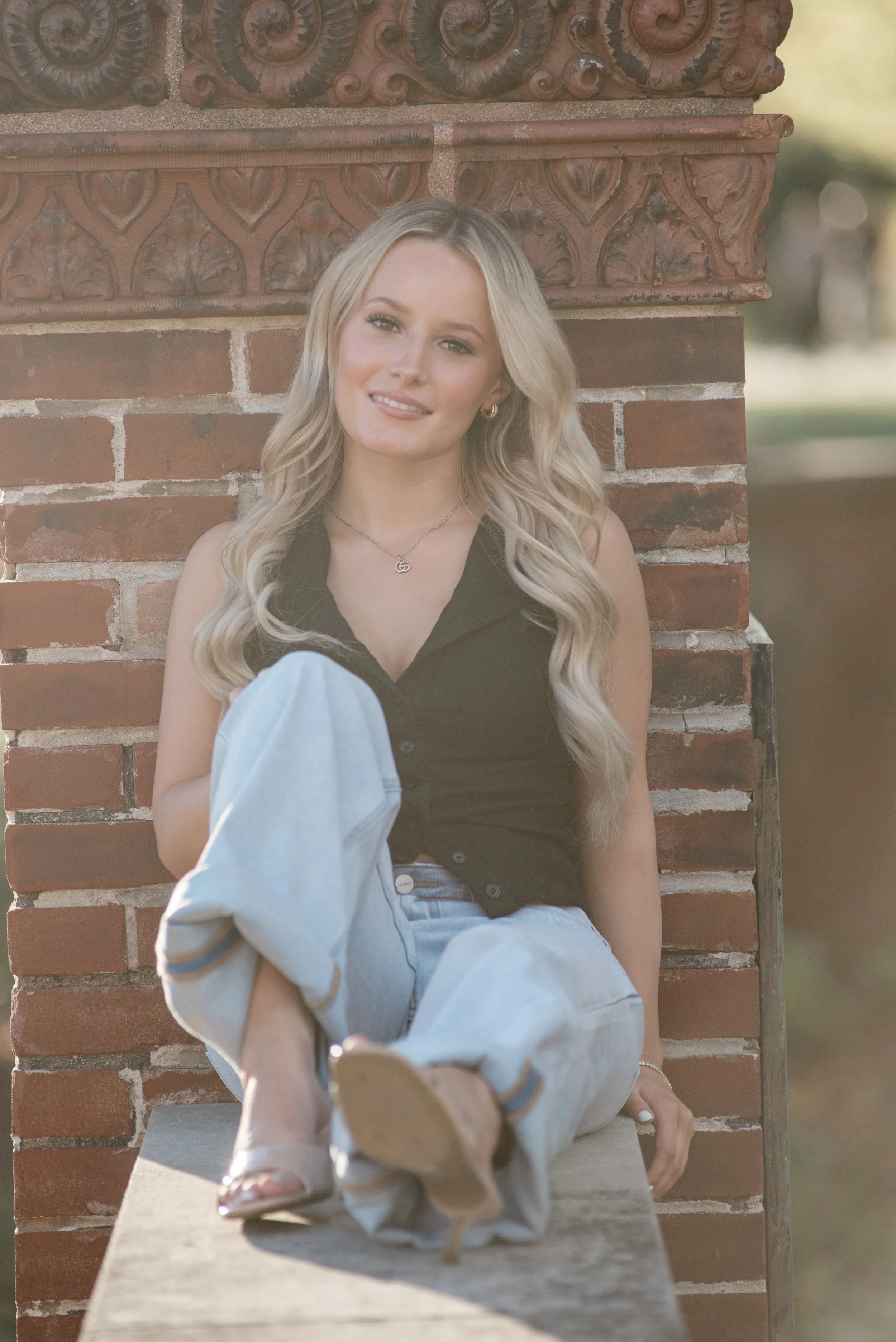 A young woman with long blonde wavy hair sits on a ledge next to a decorative brick wall, smiling at the camera. She is wearing a black sleeveless top, light blue jeans, open-toe sandals, and jewelry, including a necklace and hoop earrings. A small d
