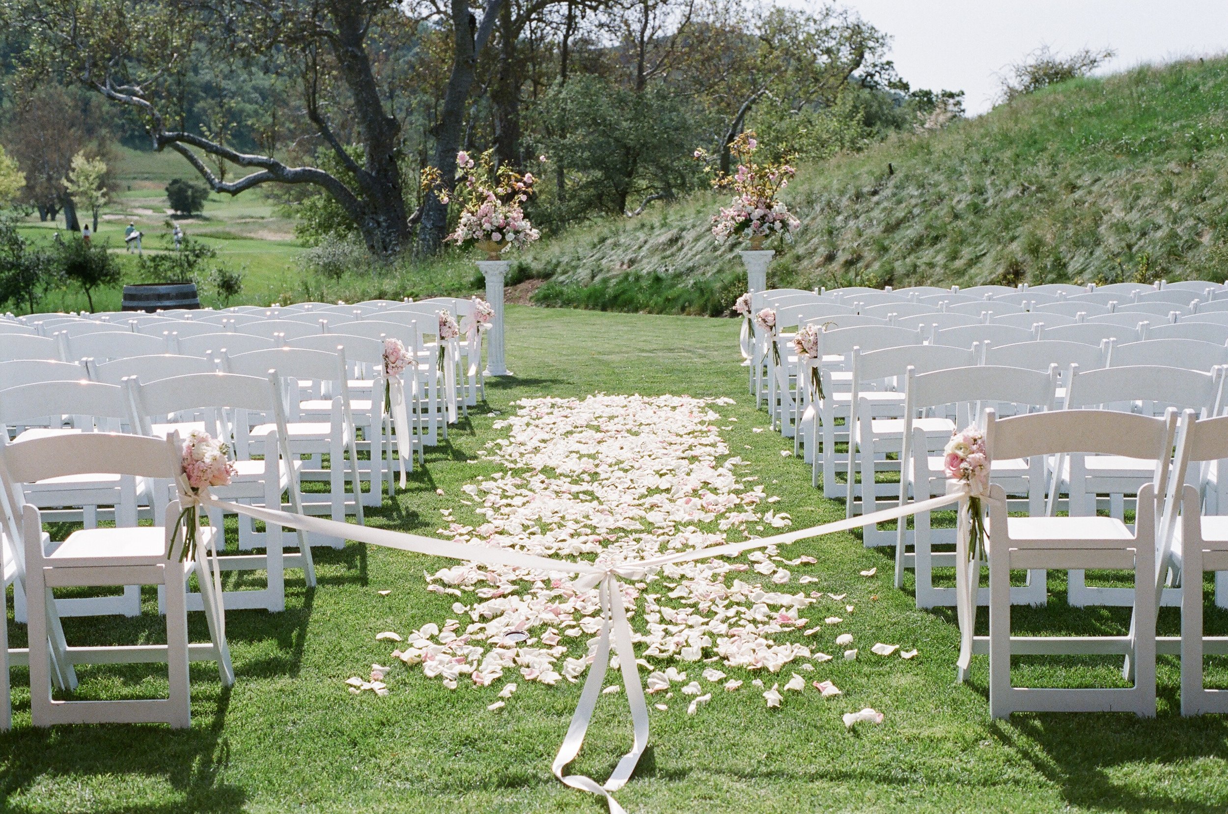 Outdoor wedding aisle lined with white chairs decorated with pink flowers, rose petals scattered along the aisle, and floral arrangements on white pedestals in a green grassy setting with trees in the background.