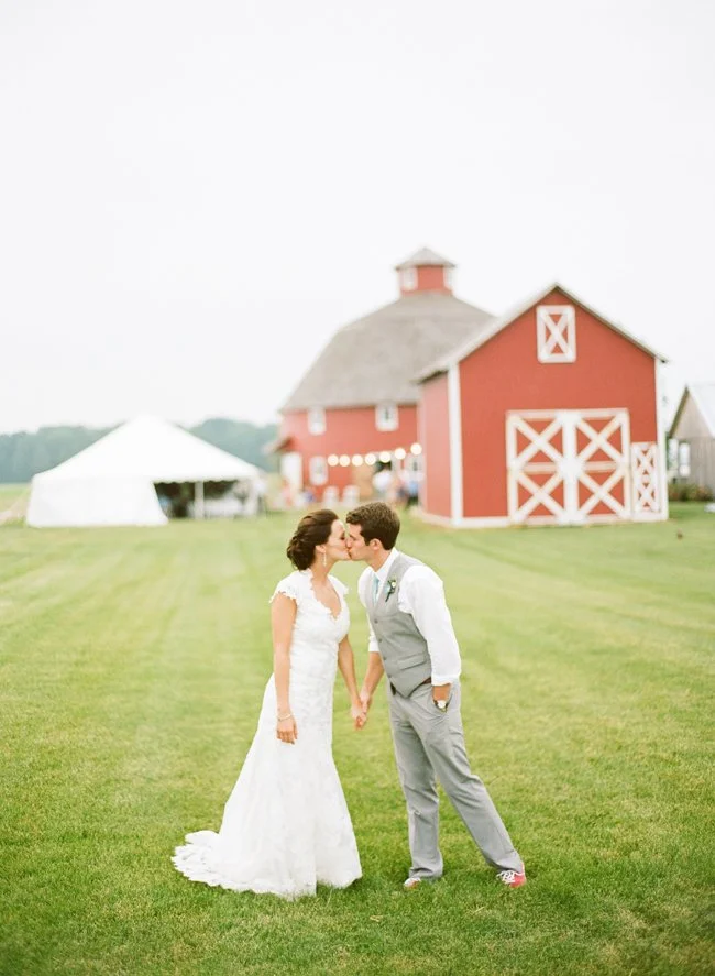 A bride and groom standing on a green lawn, holding hands, and about to kiss at their outdoor wedding, with a red barn and a white tent in the background.