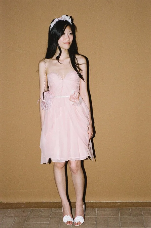 A young woman in a pink dress with lace and flower details, wearing a headband with white flowers, standing against a beige wall.