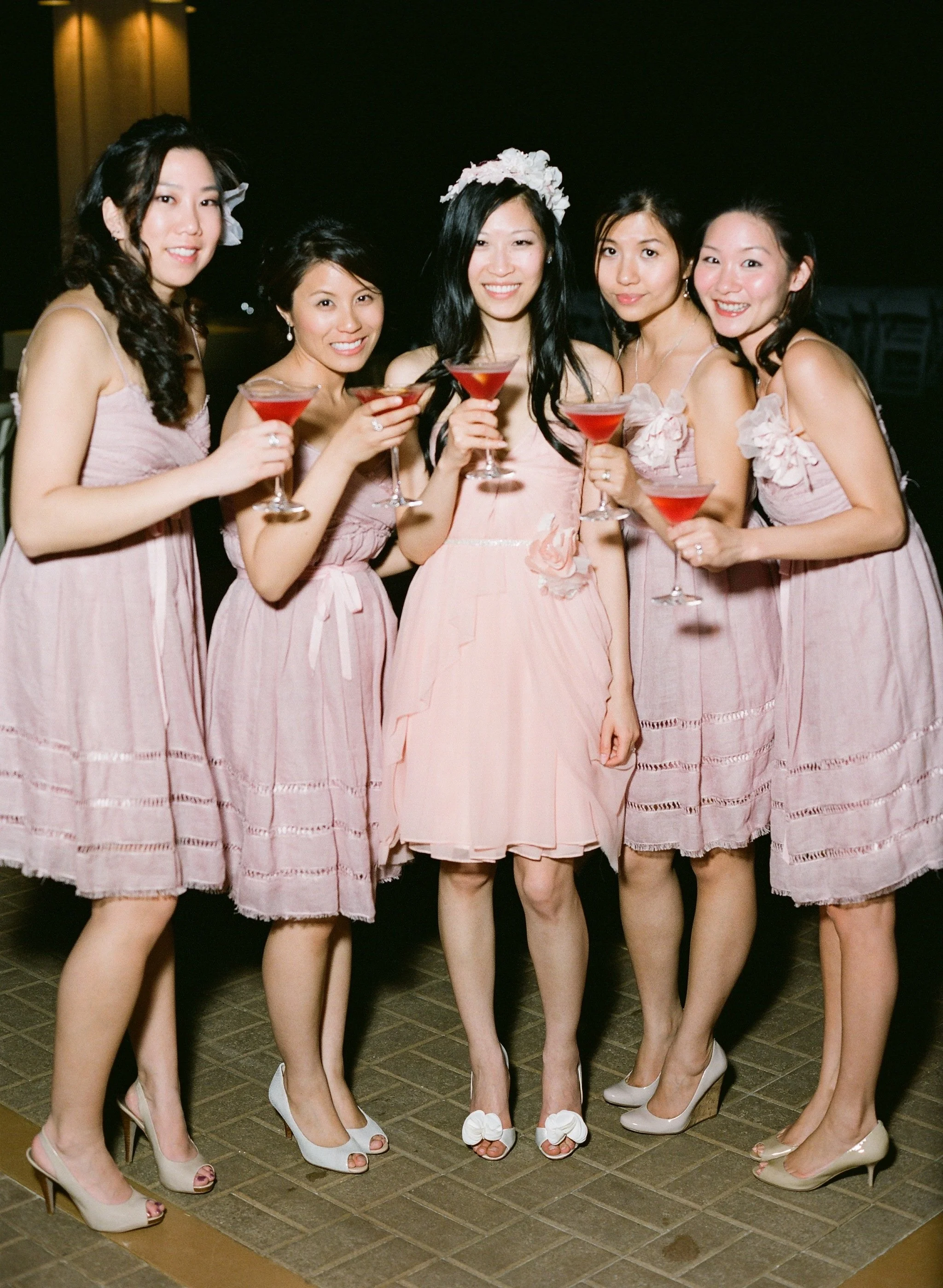 A group of five women wearing matching light pink dresses and high heels, holding pink cocktails, standing together indoors at night.