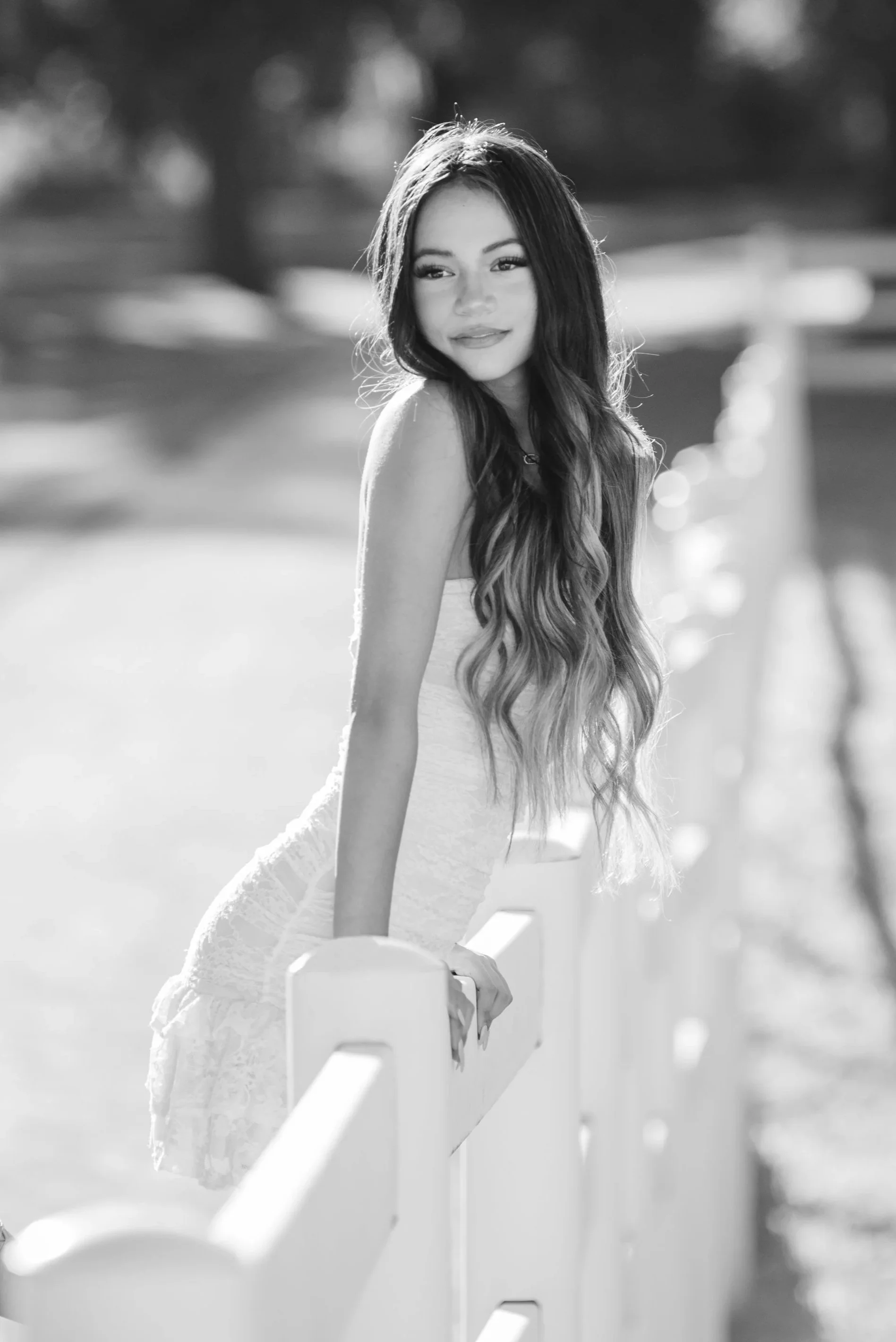 Black and white photo of a young woman with long wavy hair, looking at the camera, leaning on a white railing outdoors with blurred background.
