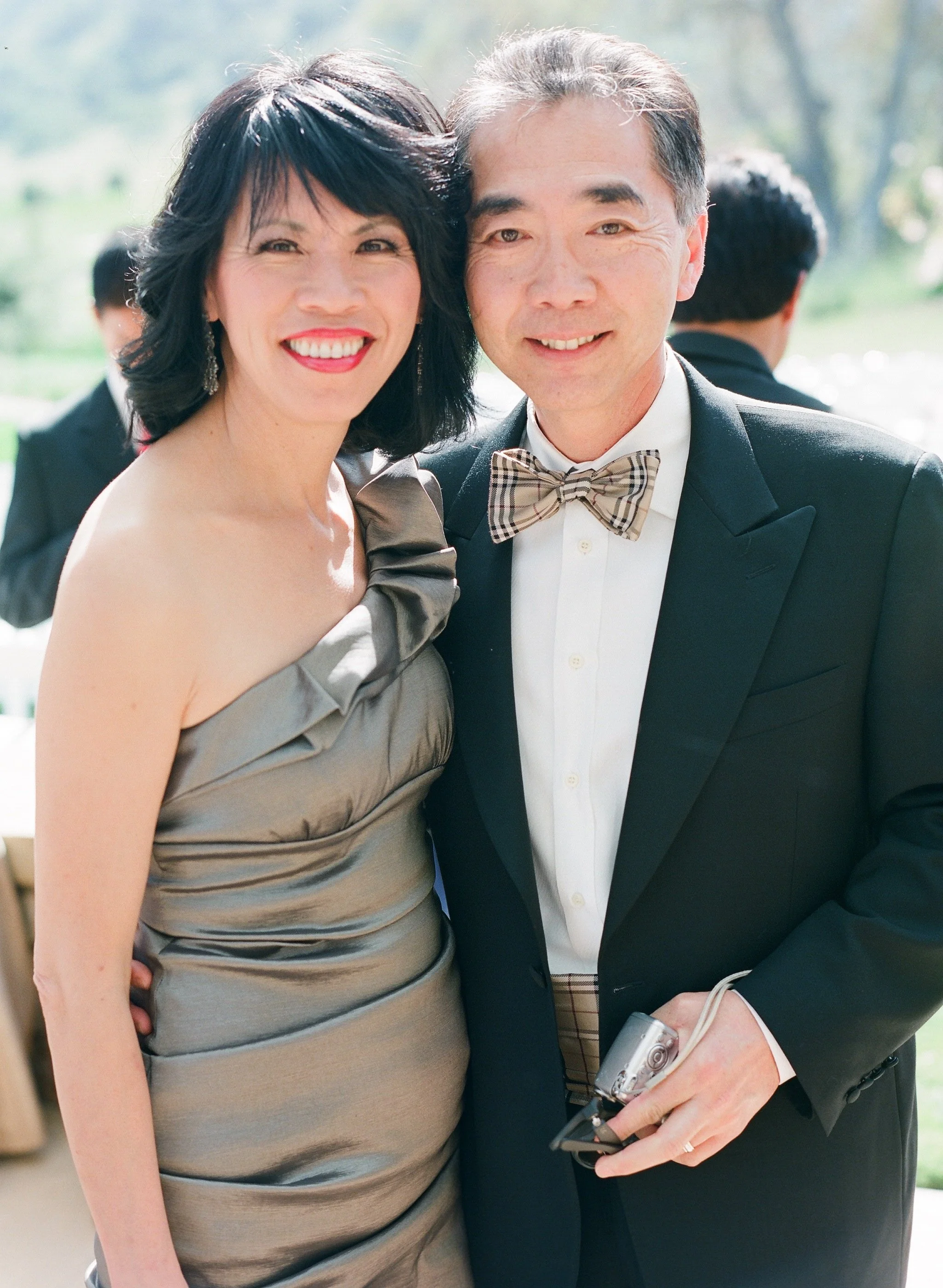 A smiling woman in a silver dress standing next to a man in a tuxedo with a plaid bow tie, outside on a sunny day, with blurred greenery in the background.