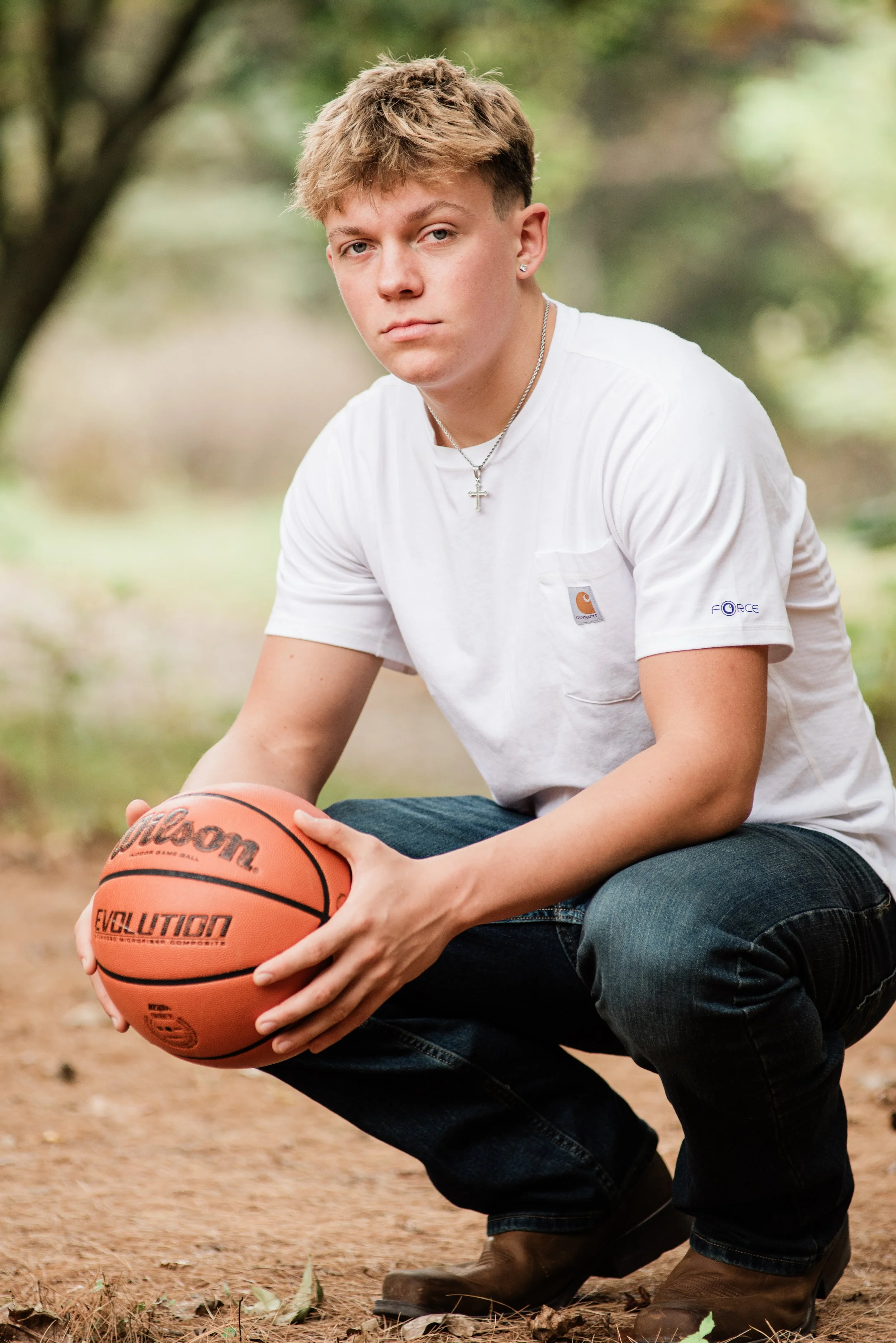 Young man crouching outdoors on dirt ground, holding a basketball, wearing a white T-shirt, jeans, and brown boots, with a background of blurred trees.