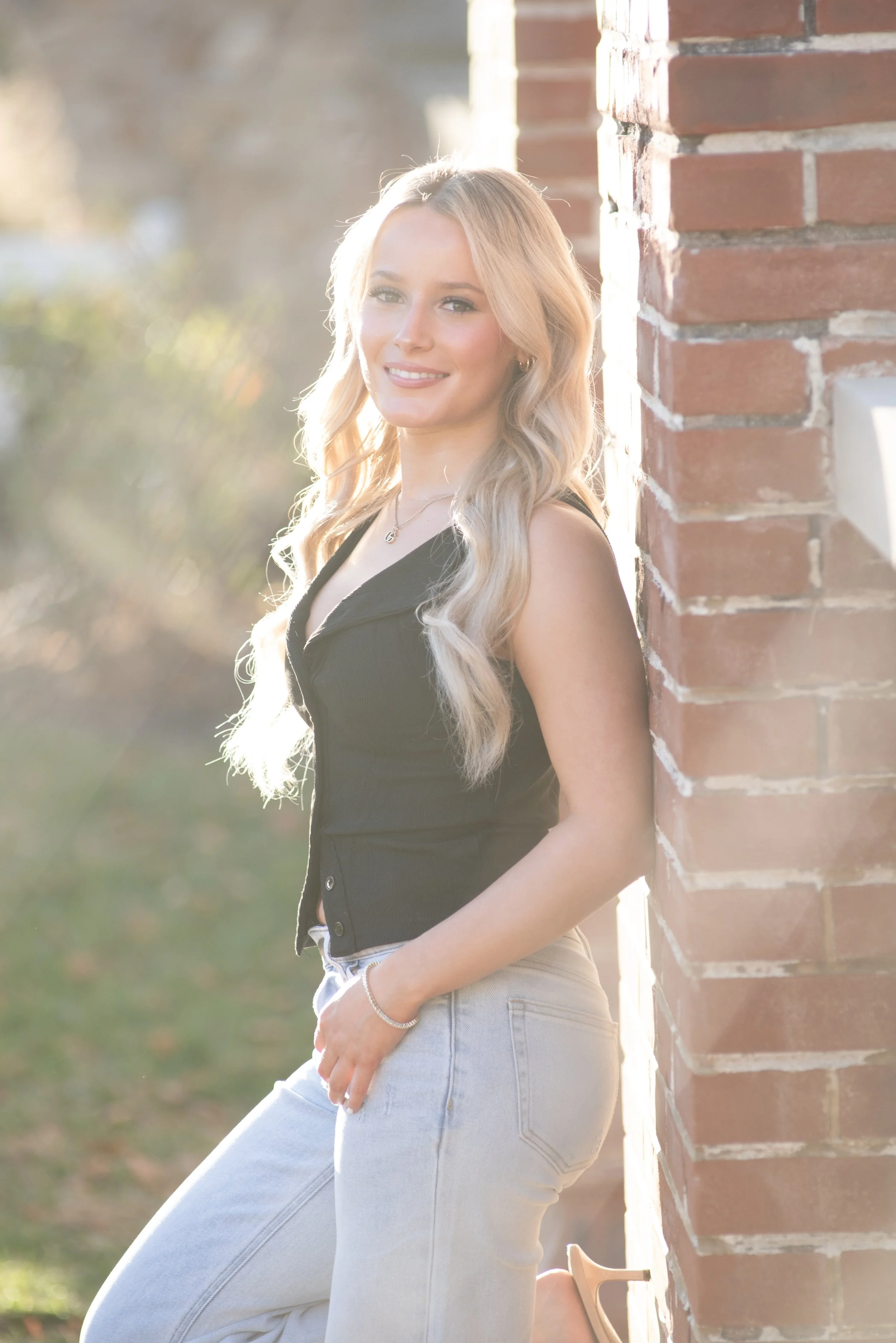 A young woman with long blond hair, wearing a black sleeveless top and light-colored jeans, standing outdoors next to a brick wall, smiling at the camera with sunlight in the background.