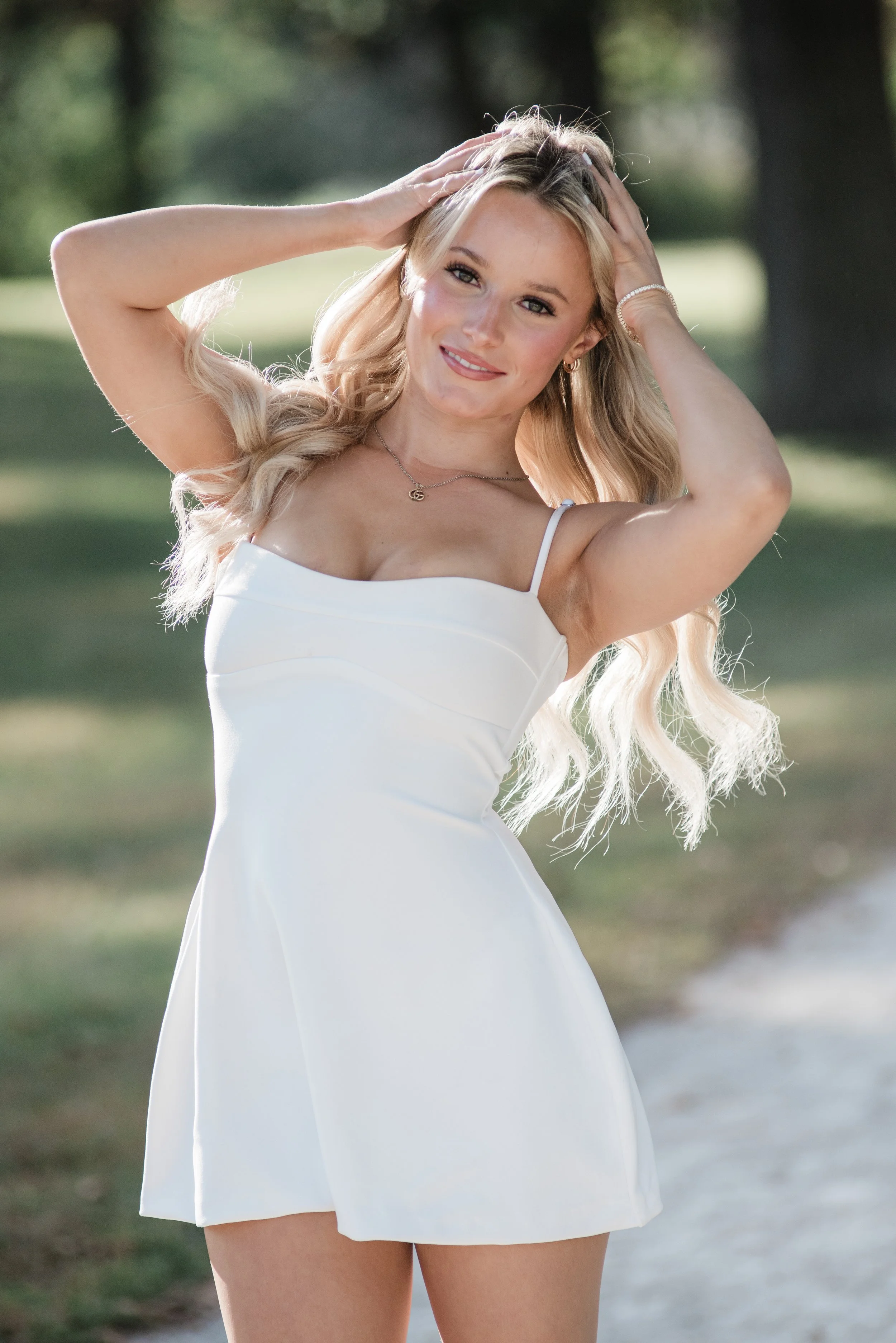 A woman with long blonde hair and fair skin smiling, holding her hands on her head, outdoors with blurred green trees and grass in the background, wearing a sleeveless white dress.